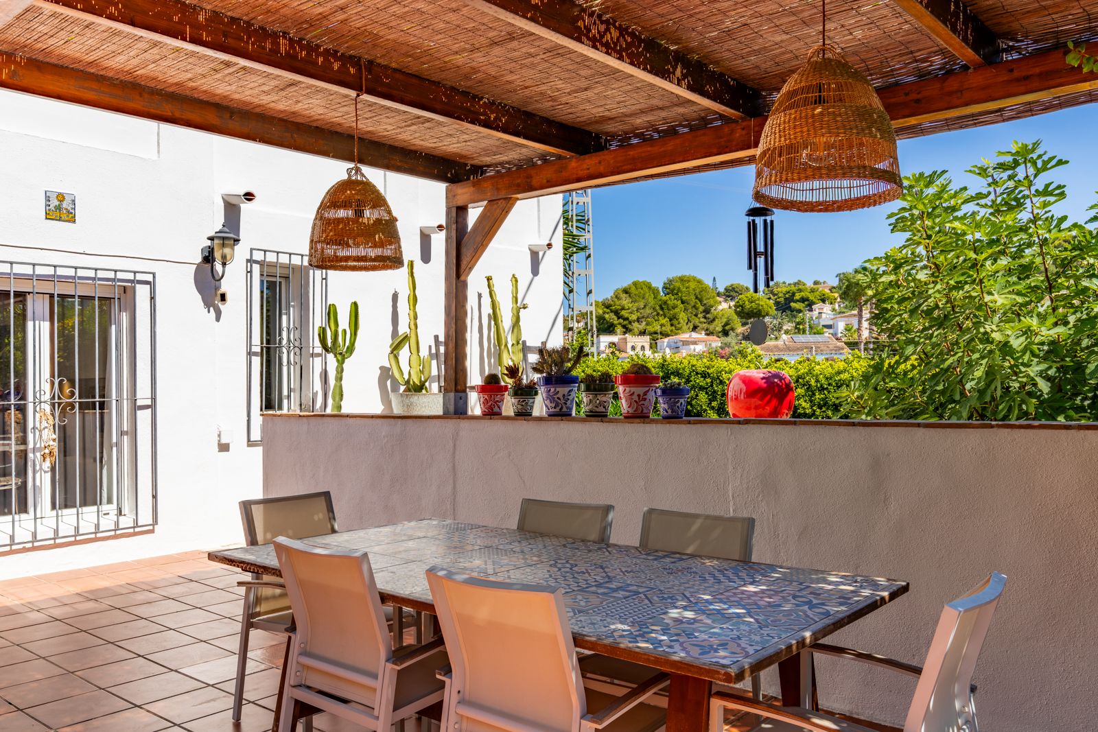 Überdachte Terrasse mit Holzpergola, dekorativem Fliesentisch und Rattanhängeleuchten, eingebettet in eine mediterrane Gartenlandschaft mit Kakteen.