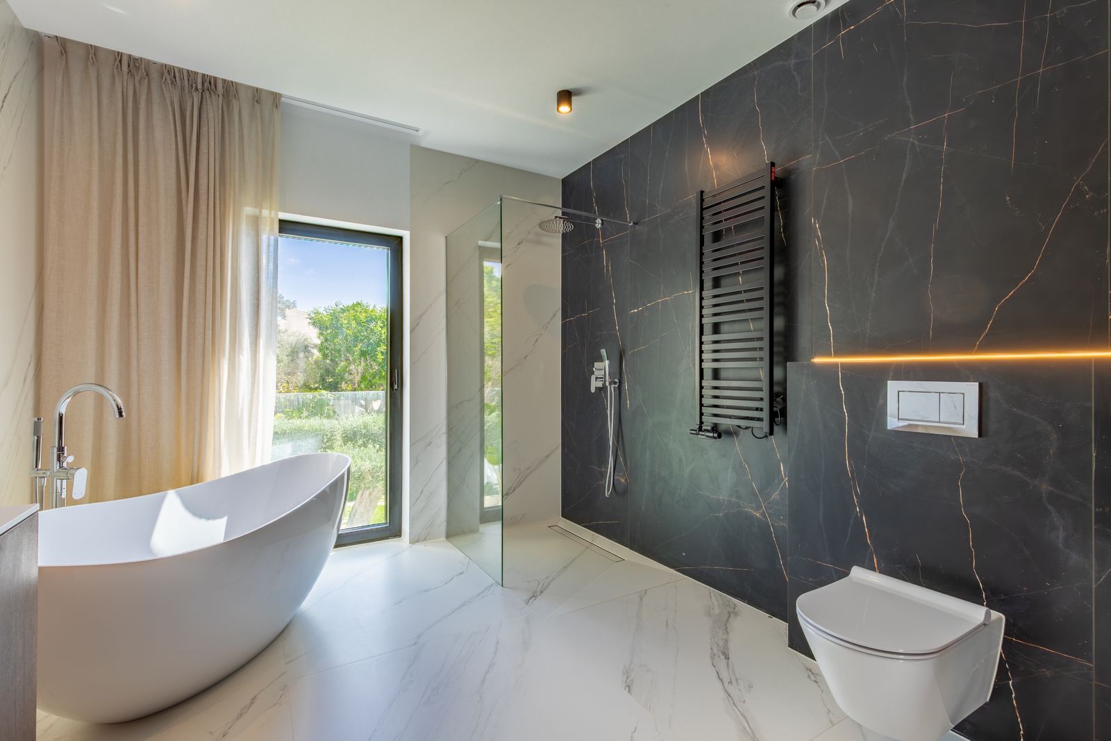 Design-Badezimmer mit freistehender Badewanne, begehbarer Dusche und kontrastierenden Feinsteinzeugwänden. Mit wandhängendem WC, schwarzem Handtuchheizkörper und Gartenblick.
