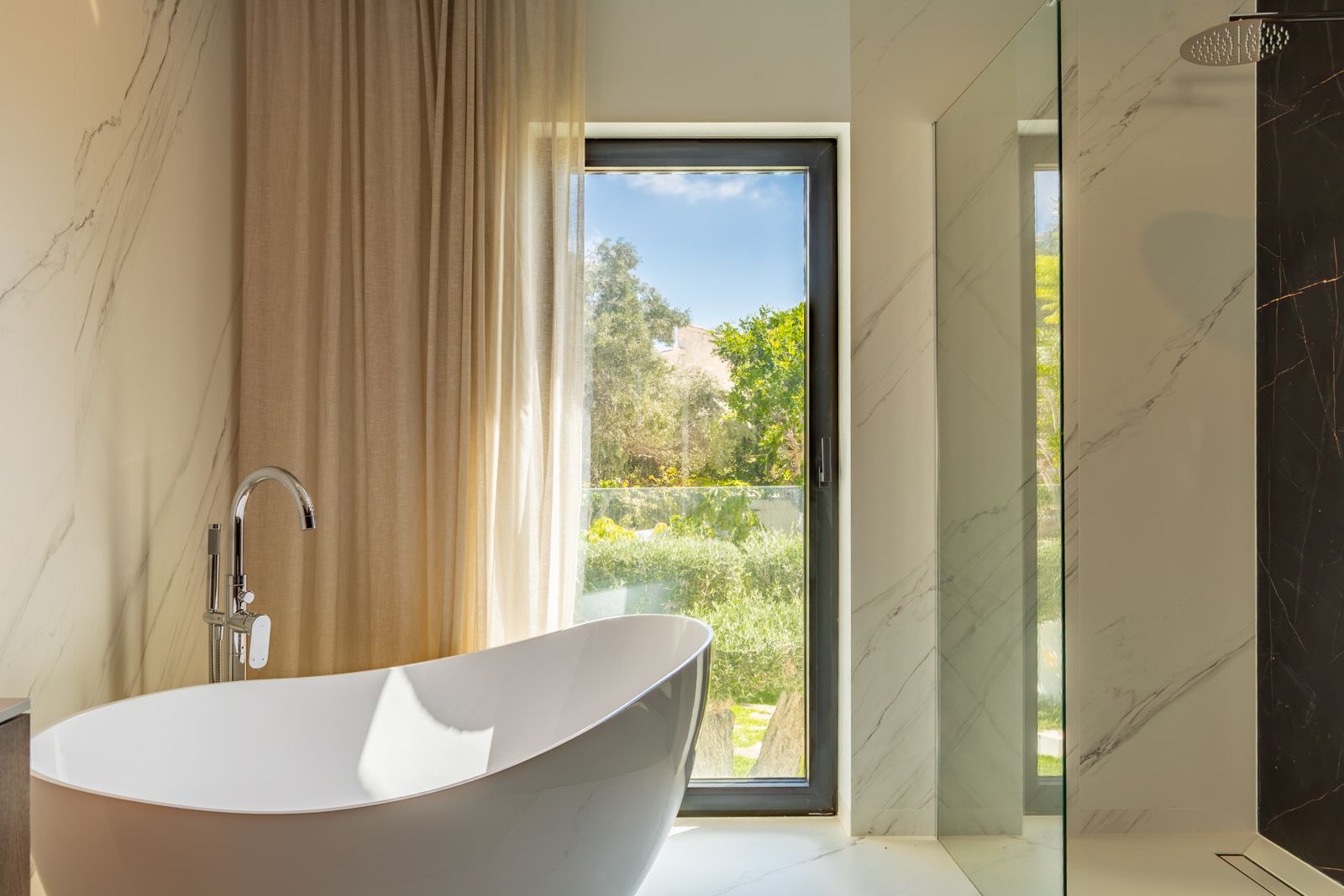 Design-Badezimmer mit freistehender Badewanne, Standarmatur und begehbarer Dusche. Marmoroptik-Feinsteinzeugwände und ein Fenster mit Blick in den mediterranen Garten.