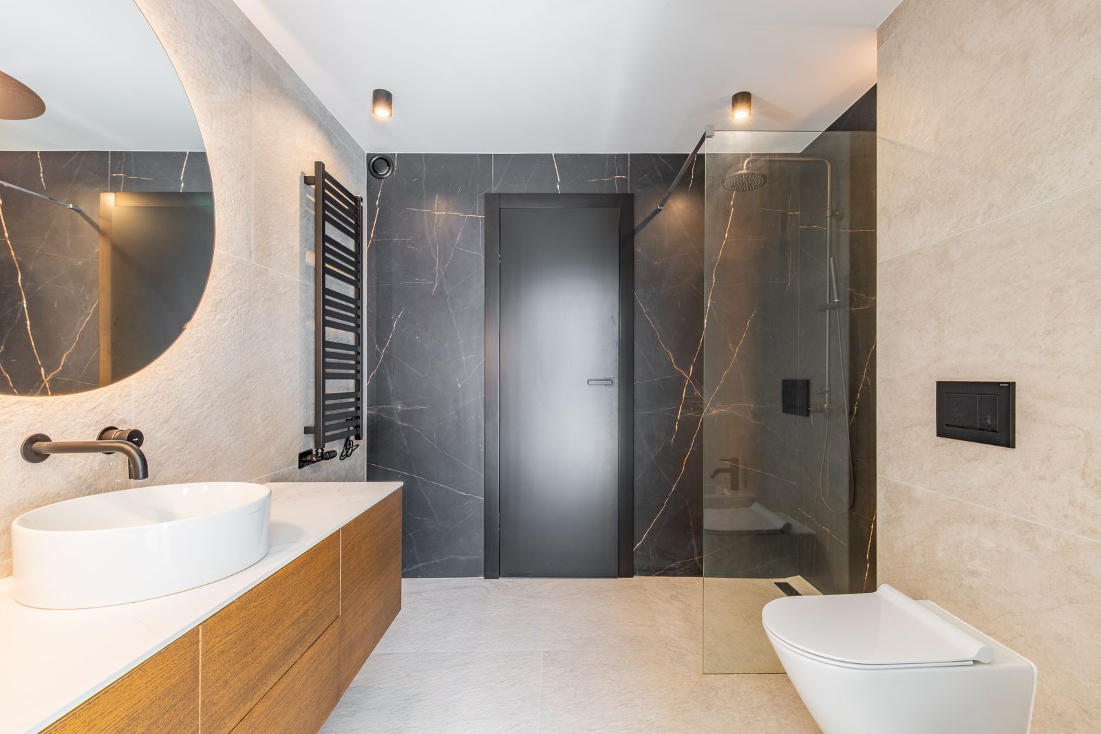 Design-Badezimmer mit großformatigem Feinsteinzeug, hinterleuchtetem Rundspiegel, Holzwaschtisch und ebenerdiger Dusche mit mattschwarzen Armaturen.