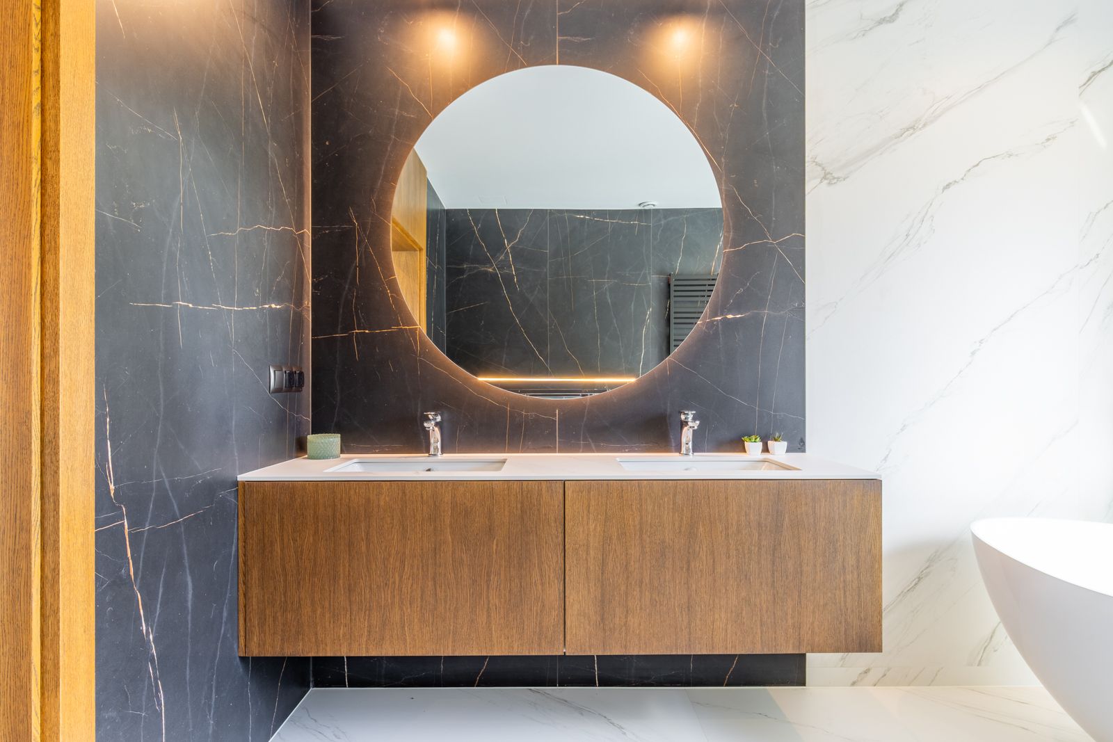 Designer-Badezimmer mit schwebendem Waschtisch aus Eichenholz, Doppelwaschbecken und hinterleuchtetem Rundspiegel vor dunklen Feinsteinzeugfliesen mit Goldmaserung.
