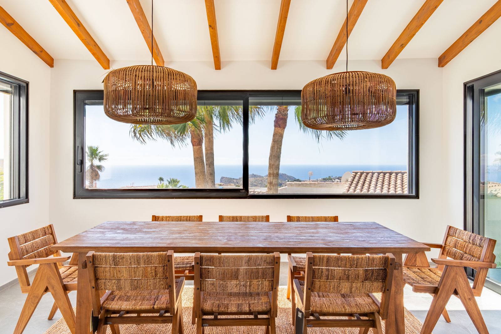 Essbereich mit Holzbalkendecke, Massivholztisch und Rattanleuchten. Das große Fenster bietet einen Panoramablick auf das Mittelmeer und die Palmen.