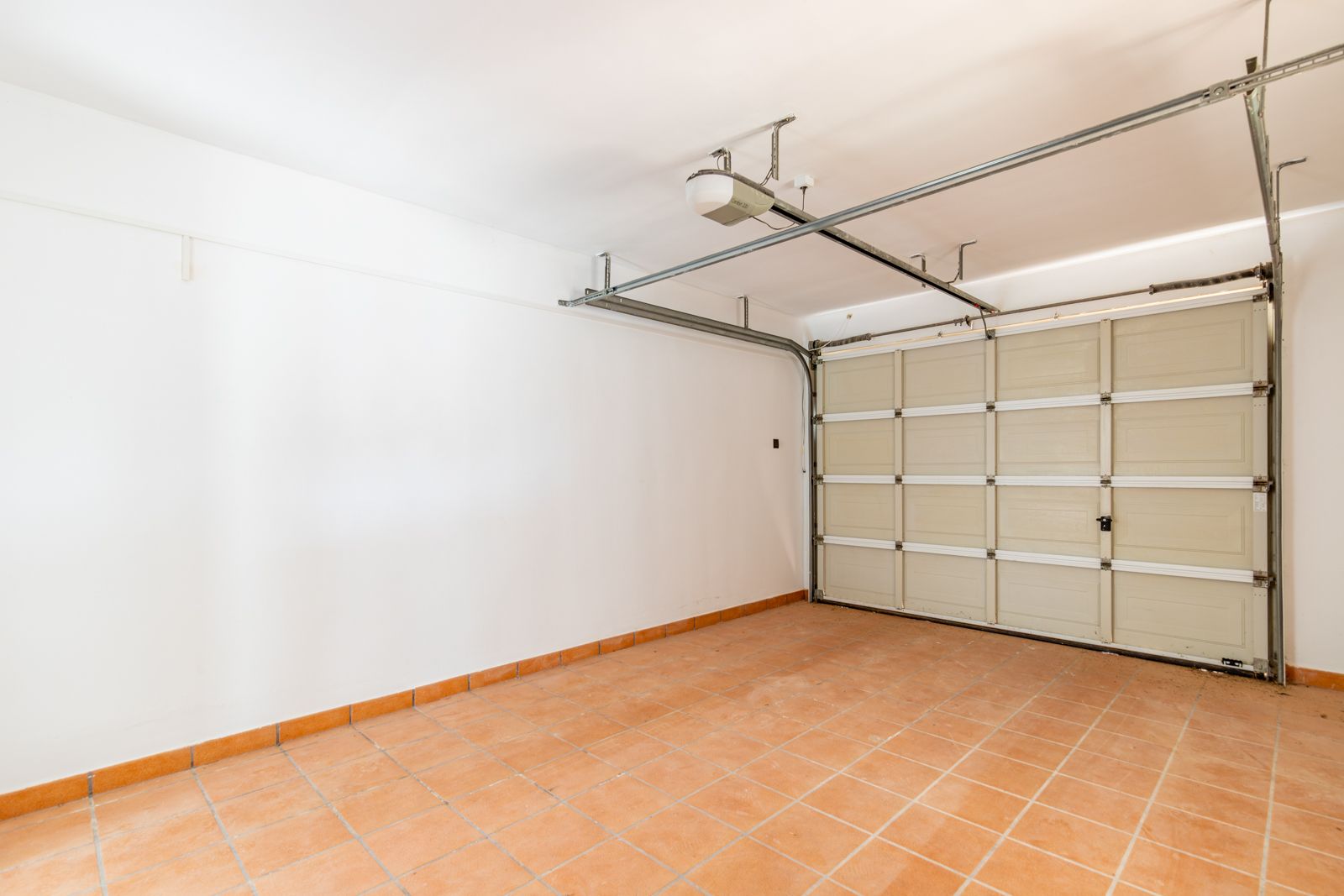 Geschlossene Garage mit terracottafarbenen Bodenfliesen, glatten weißen Wänden und einem motorisierten Sektionaltor mit automatischem Deckenantrieb.