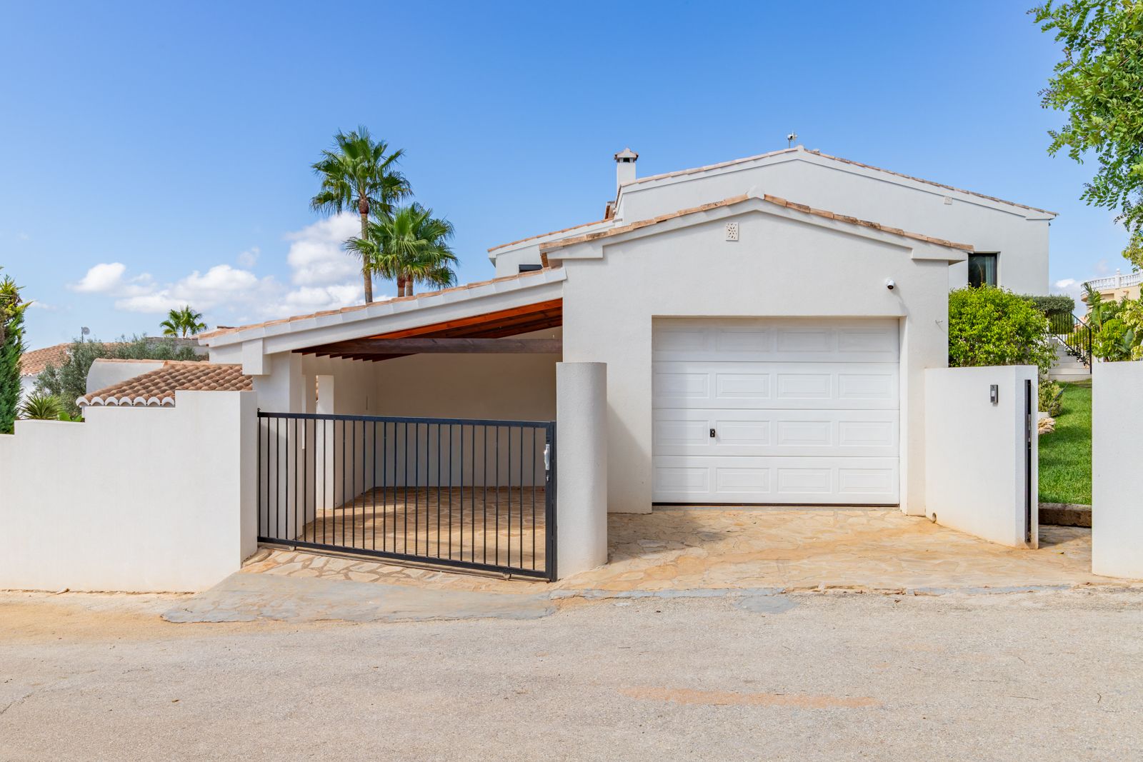 Haupteingang mit geschlossener Garage und überdachtem Carport. Natursteinpflaster, ein schwarzes Metalltor und weiße mediterrane Architektur prägen den Außenbereich.