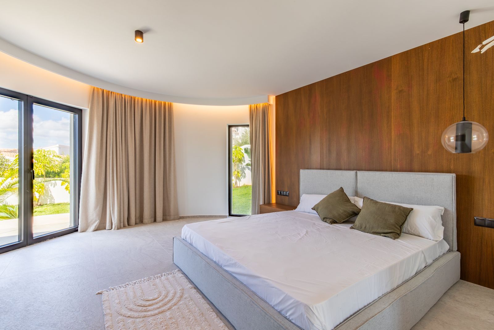 Hauptschlafzimmer mit Wandverkleidung aus Natureiche, geschwungener Decke mit indirekter LED-Beleuchtung und direktem Gartenzugang durch große Glasfronten.