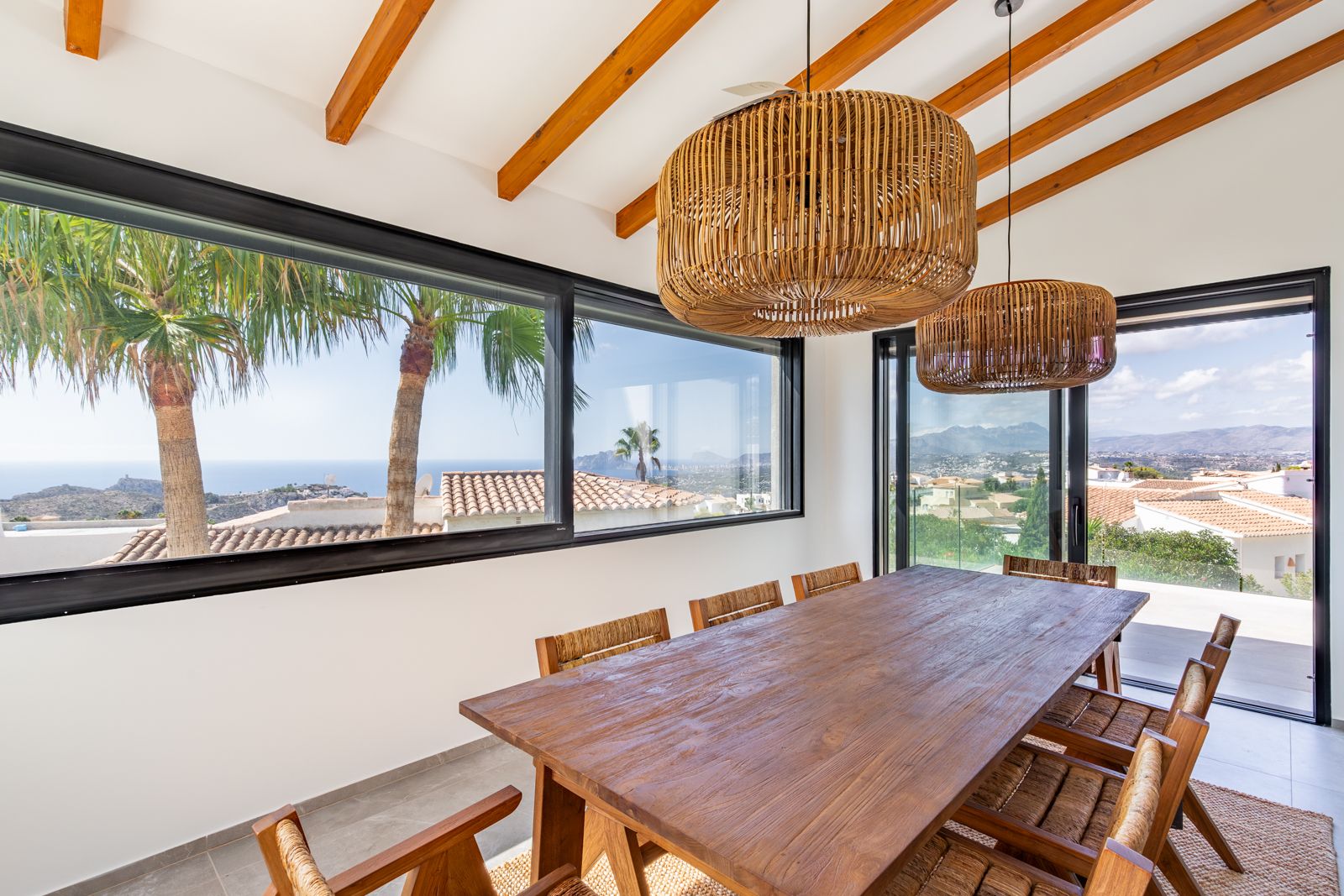 Heller Essbereich mit Sichtbalken, massivem Holztisch und Rattan-Pendelleuchten. Große Fensterfronten bieten einen Panoramablick auf das Mittelmeer und die Berge.