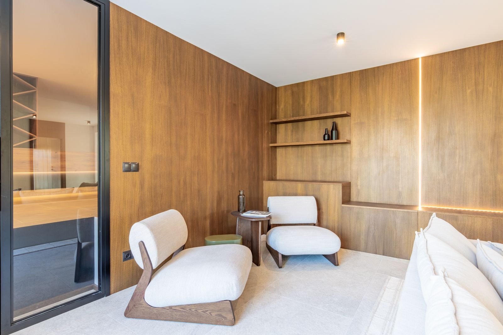 Loungebereich mit Wandverkleidung aus Eichenfurnier, integrierter LED-Beleuchtung und großformatigen Feinsteinzeugfliesen. Design-Sessel in weißer Polsterung.