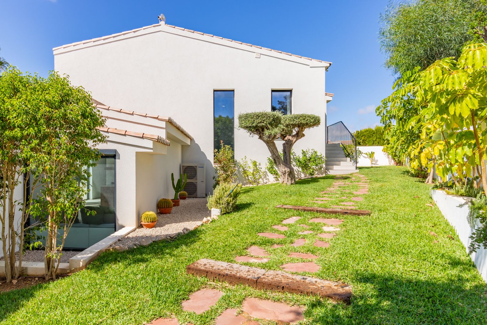 Mediterraner Garten mit gepflegtem Rasen, dekorativem Olivenbaum und Natursteinpfad, der zur modernen weißen Fassade der Villa führt.