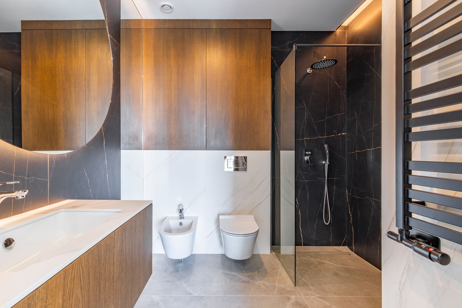 Modernes Badezimmer mit dunklen Feinsteinzeugfliesen, begehbarer Dusche, wandhängenden Sanitärobjekten und Eichenmöbeln unter einem großen, beleuchteten Rundspiegel.