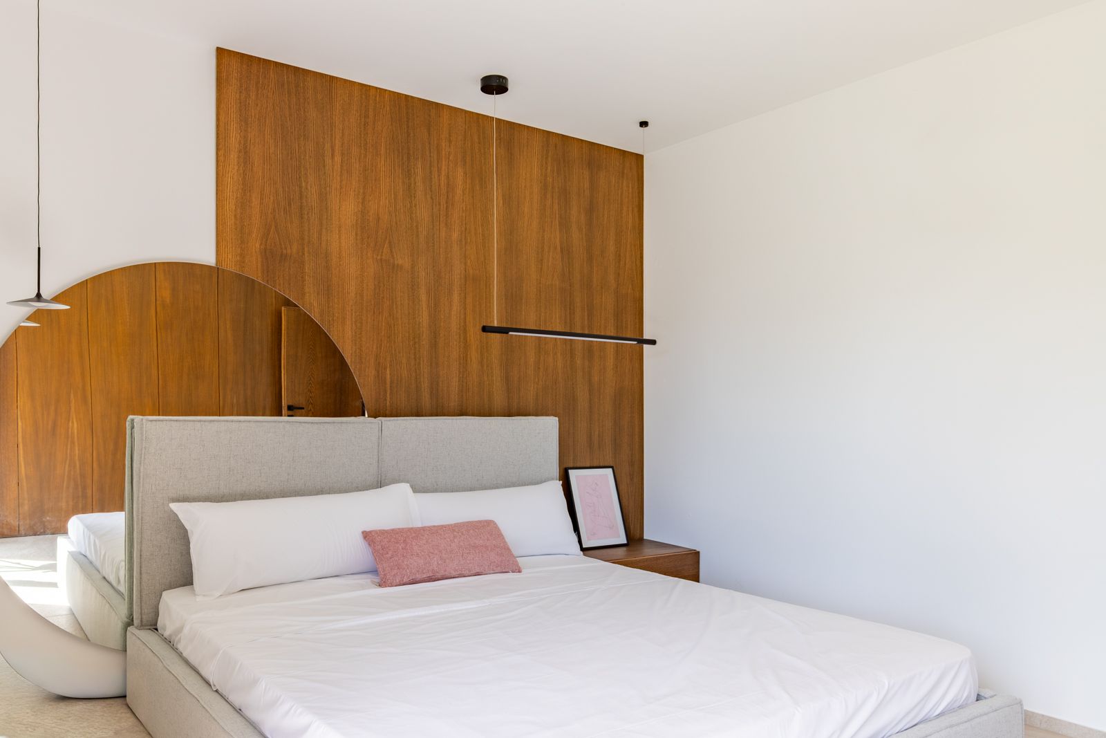 Modernes Schlafzimmer mit Wandpaneelen aus Natureiche, grauem Textil-Kopfteil und großem Rundspiegel. Minimalistische schwarze Pendelleuchten und integrierter Nachttisch.