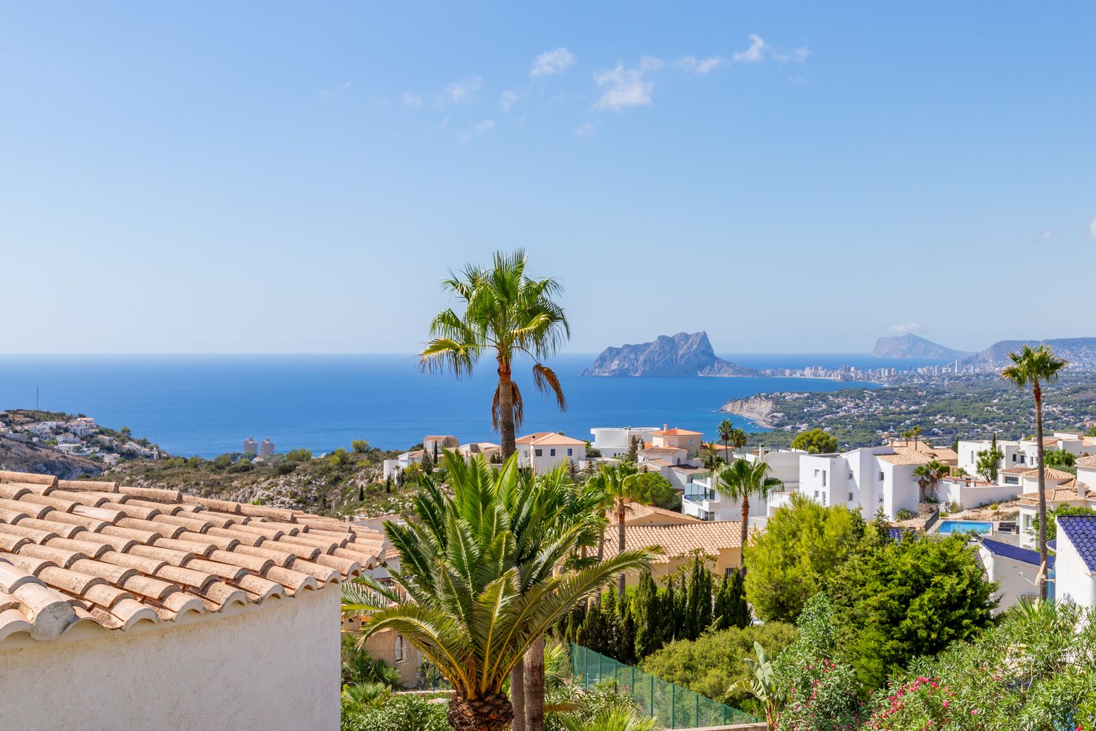 Panoramablick auf das Mittelmeer und den Peñón de Ifach, mit Küstenarchitektur und üppigen Palmen unter strahlend blauem Himmel.