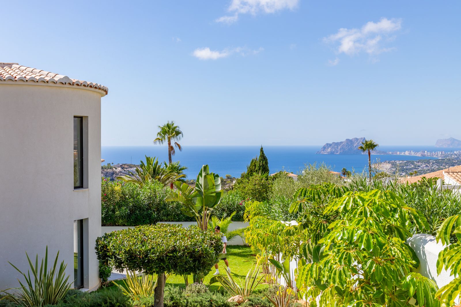 Panoramablick auf das Mittelmeer und den Peñón de Ifach vom mediterranen Garten aus, der die moderne, geschwungene Fassade der Villa umgibt.
