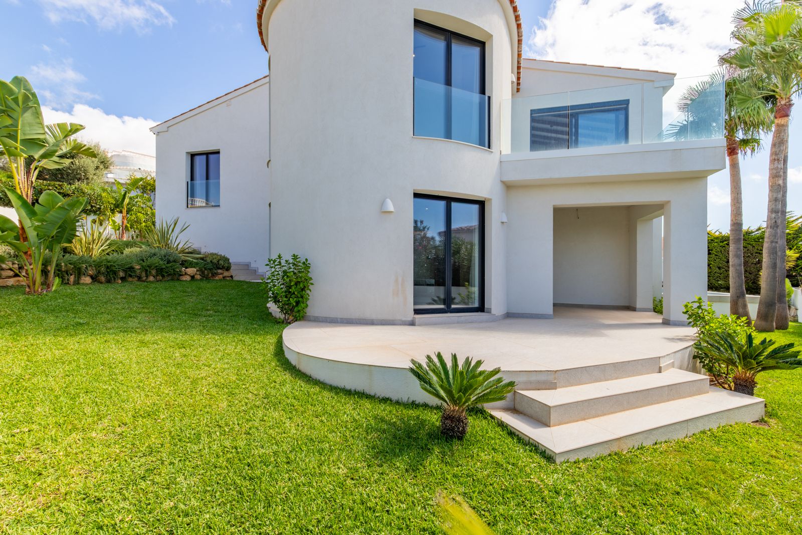 Rückansicht der Villa mit geschwungenem Turm, dunklen Aluminiumfenstern und Glasgeländern. Der gepflegte Garten bietet Rasenflächen und mediterrane Bepflanzung.