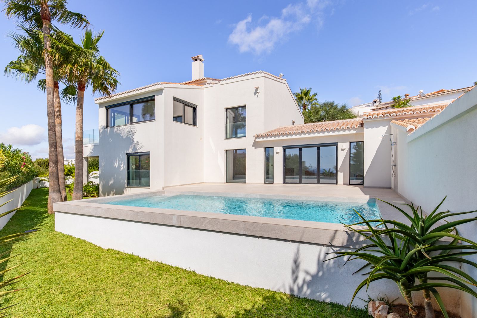 Rückansicht der Villa mit Salzwasserpool, Terrasse aus Feinsteinzeug und dreifach verglasten Fenstern. Gartenanlage mit Rasen und Palmen.
