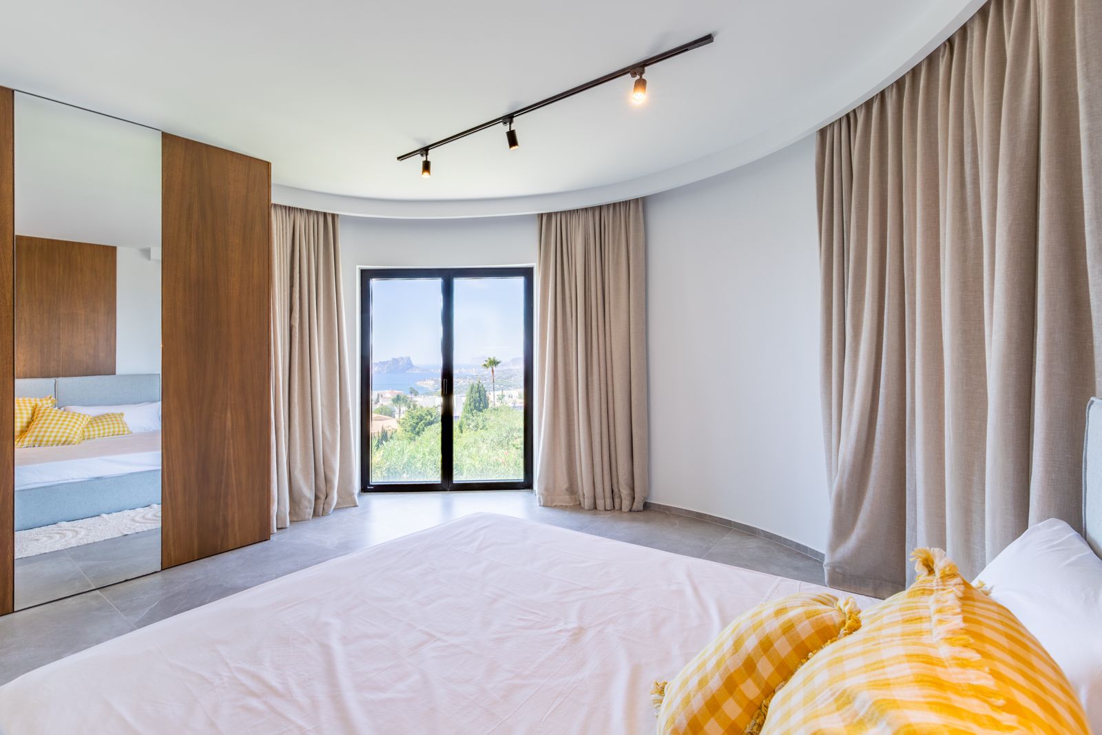 Schlafzimmer mit geschwungener Wand, maßgefertigtem Eichenschrank und bodentiefen Fenstern, die einen weiten Blick auf die Küste von Moraira bieten.