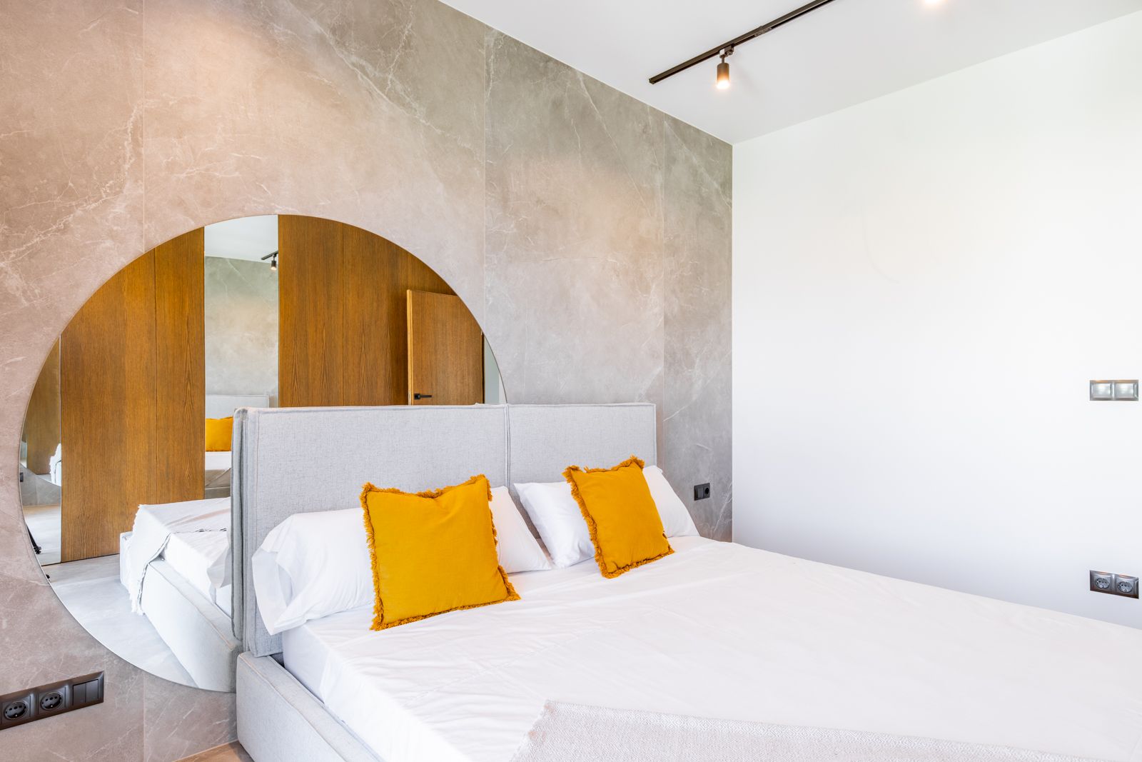 Schlafzimmer mit großformatiger Feinsteinzeugwand und integriertem Rundspiegel. Mit gepolstertem Kopfteil, Eichenholzverkleidung und moderner Schienenbeleuchtung.
