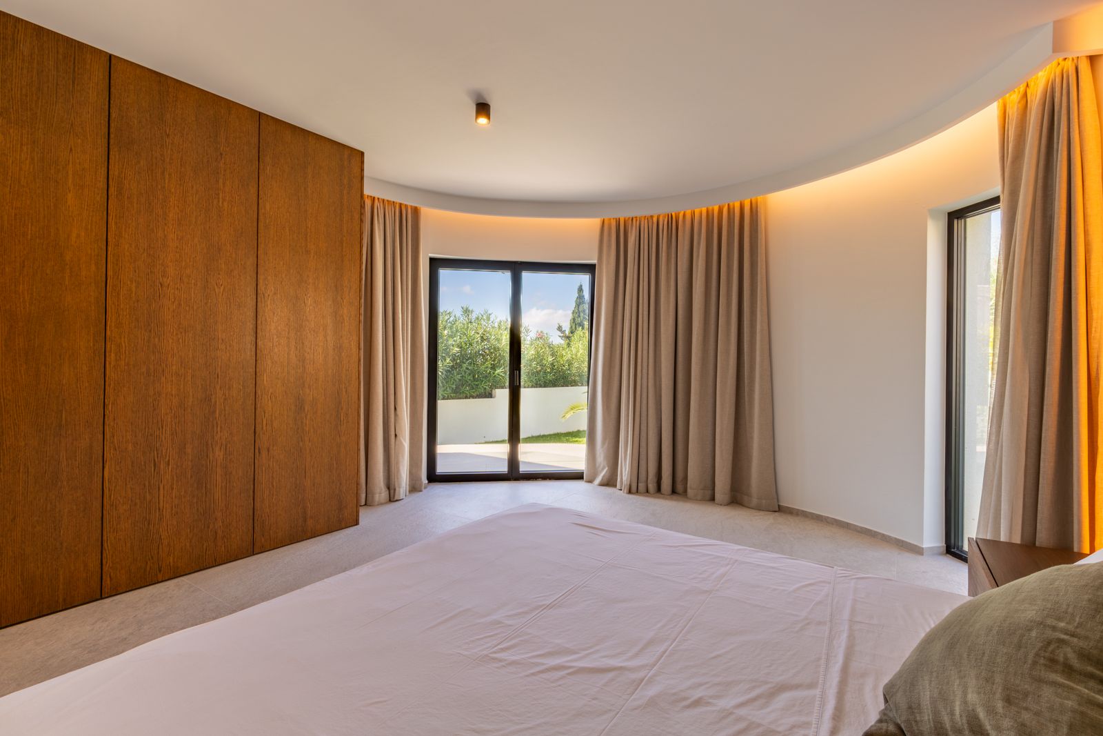 Schlafzimmer mit kurviger Architektur, Einbauschränken aus Eichenholz und indirektem Licht. Bodentiefe Fenster ermöglichen den direkten Zugang zum Garten.