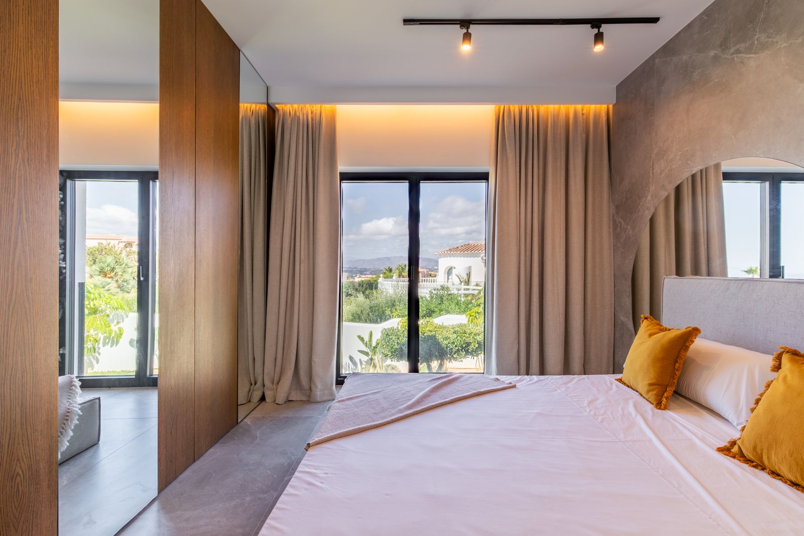 Schlafzimmer mit Wandverkleidung aus Eiche, Feinsteinzeugböden und großen Fenstern mit Blick auf den mediterranen Garten und die Landschaft.