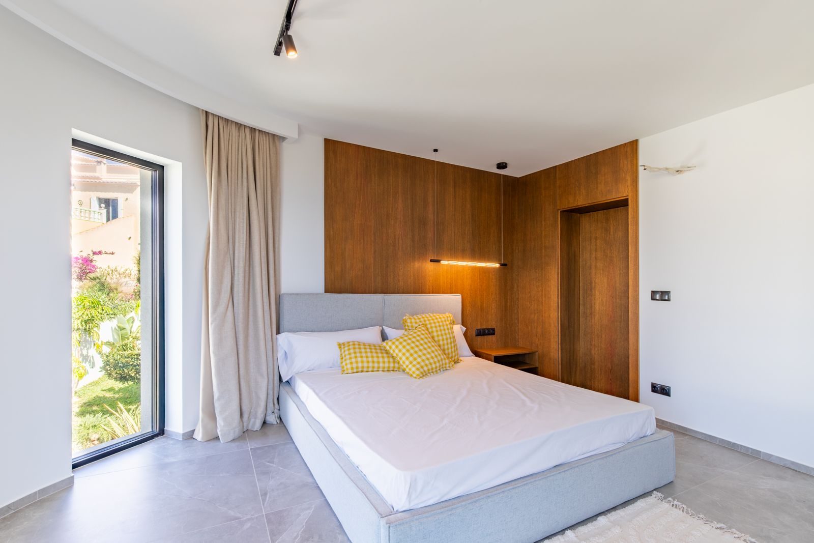 Schlafzimmer mit Wandverkleidung aus Eichenholz, Designer-Pendelleuchte und großformatigen Feinsteinzeugfliesen. Bodentiefe Fenster bieten direkten Blick in den Garten.