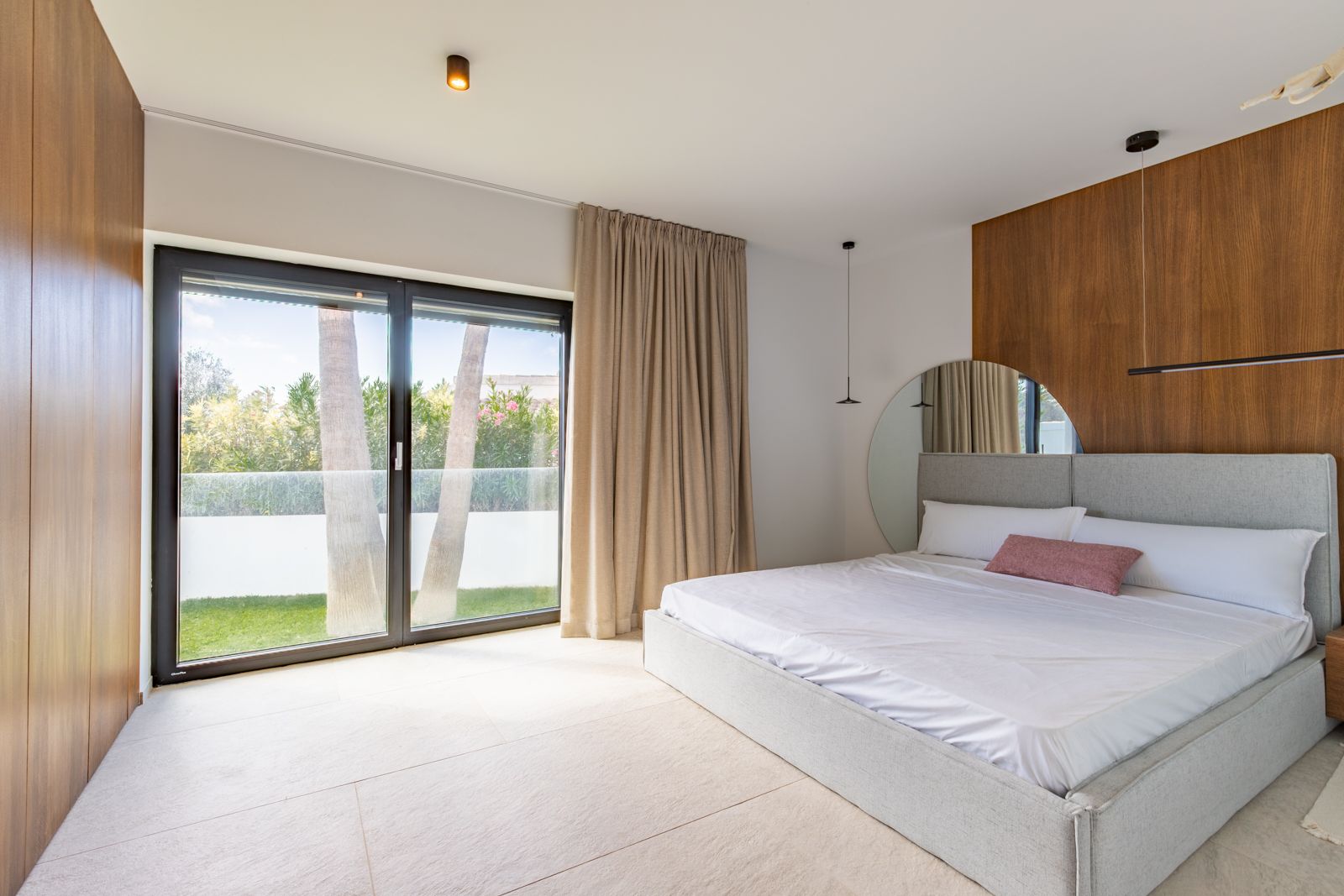 Schlafzimmer mit Wandverkleidung aus Natureiche, großformatigem Feinsteinzeugboden und direktem Gartenzugang durch dreifach verglaste Schiebetüren.