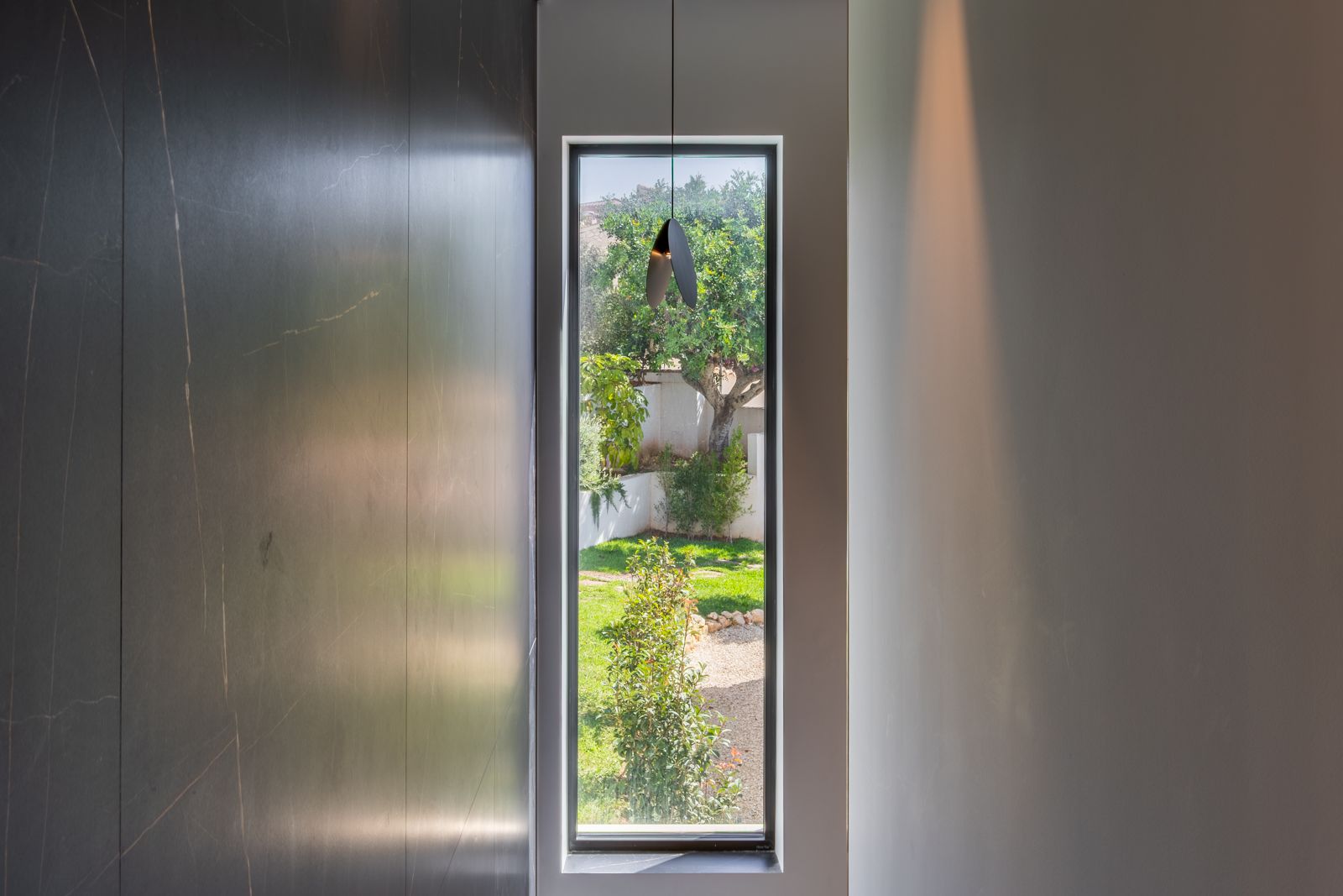 Vertikales dreifach verglastes Fenster mit Blick auf den Garten, flankiert von großformatigen dunklen Feinsteinzeugplatten und einer minimalistischen Designerleuchte.