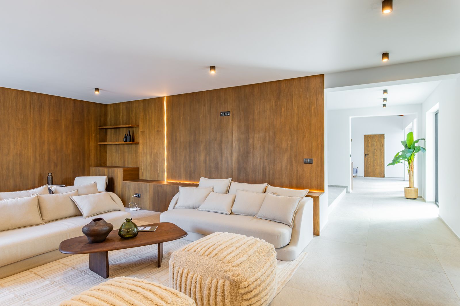 Wohnbereich mit Wandpaneelen aus Natureiche und LED-Beleuchtung. Neutrale Sofas auf großformatigem Feinsteinzeugboden und technische Deckenstrahler.