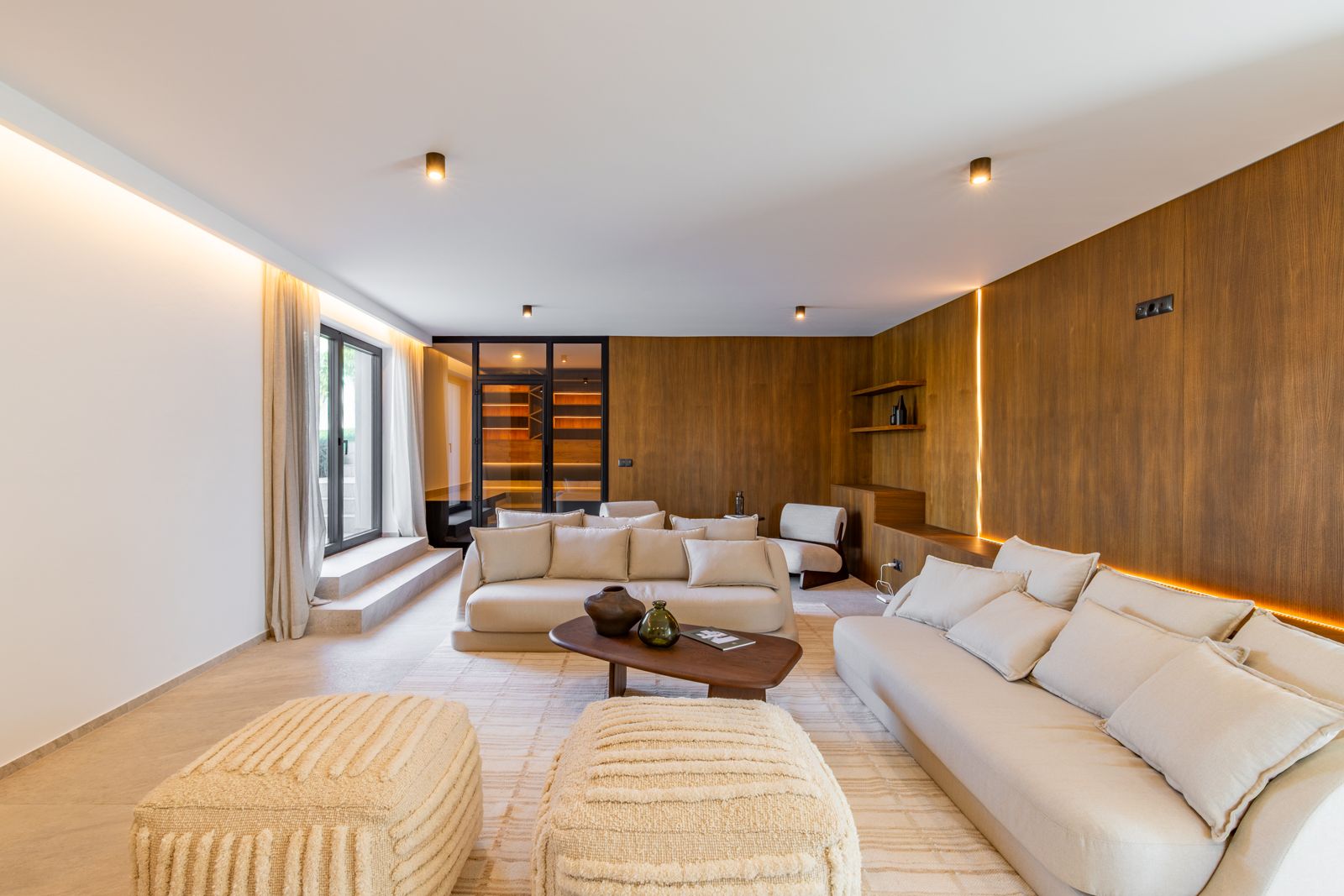 Wohnbereich mit Wandverkleidung aus Eichenholz und integrierter LED-Beleuchtung. Neutrale Sofas, Feinsteinzeugböden und Blick auf einen verglasten Weinkeller.