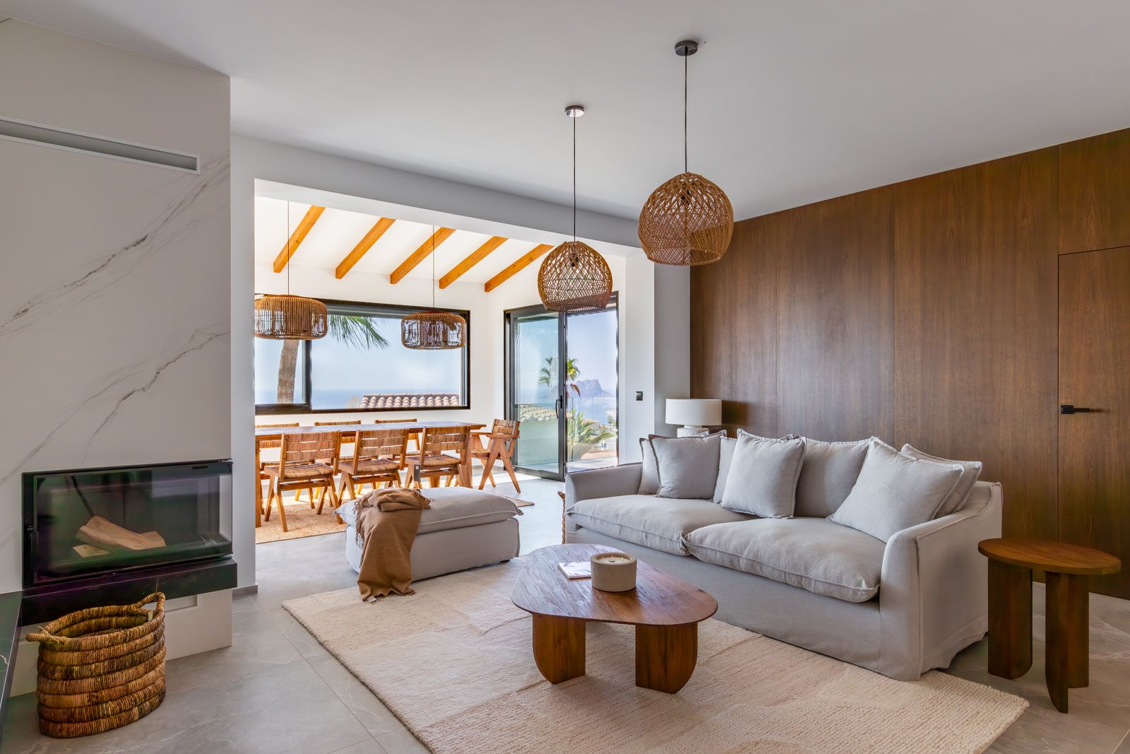 Wohnzimmer mit integriertem Kamin, Eichenholz-Wandpaneelen und Meerblick. Akzentuiert durch Rattanhängeleuchten und einen Essbereich mit sichtbaren Holzbalken.