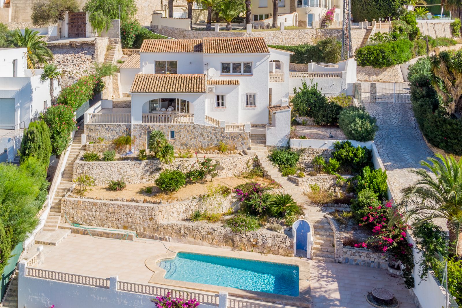 Außenansicht der Villa mit weißen Fassaden, Terrakottadächern und terrassierten mediterranen Gärten mit Natursteinmauern, die zum Pool führen.