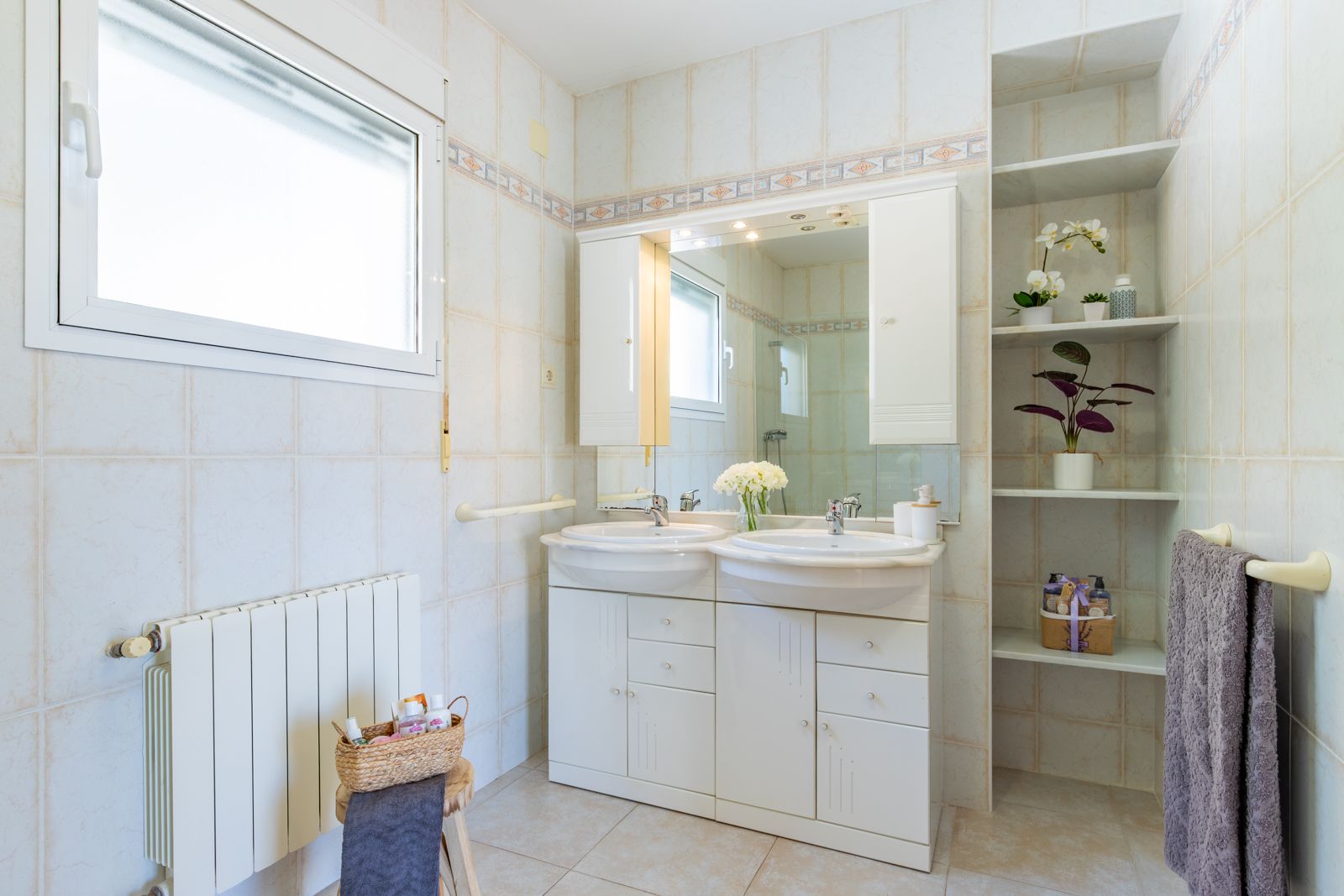 Badezimmer mit Doppelwaschtisch, weißen Unterschränken, beleuchtetem Spiegel und Einbauregalen. Geflieste Wände mit Zierborte und ein wandmontierter Heizkörper.