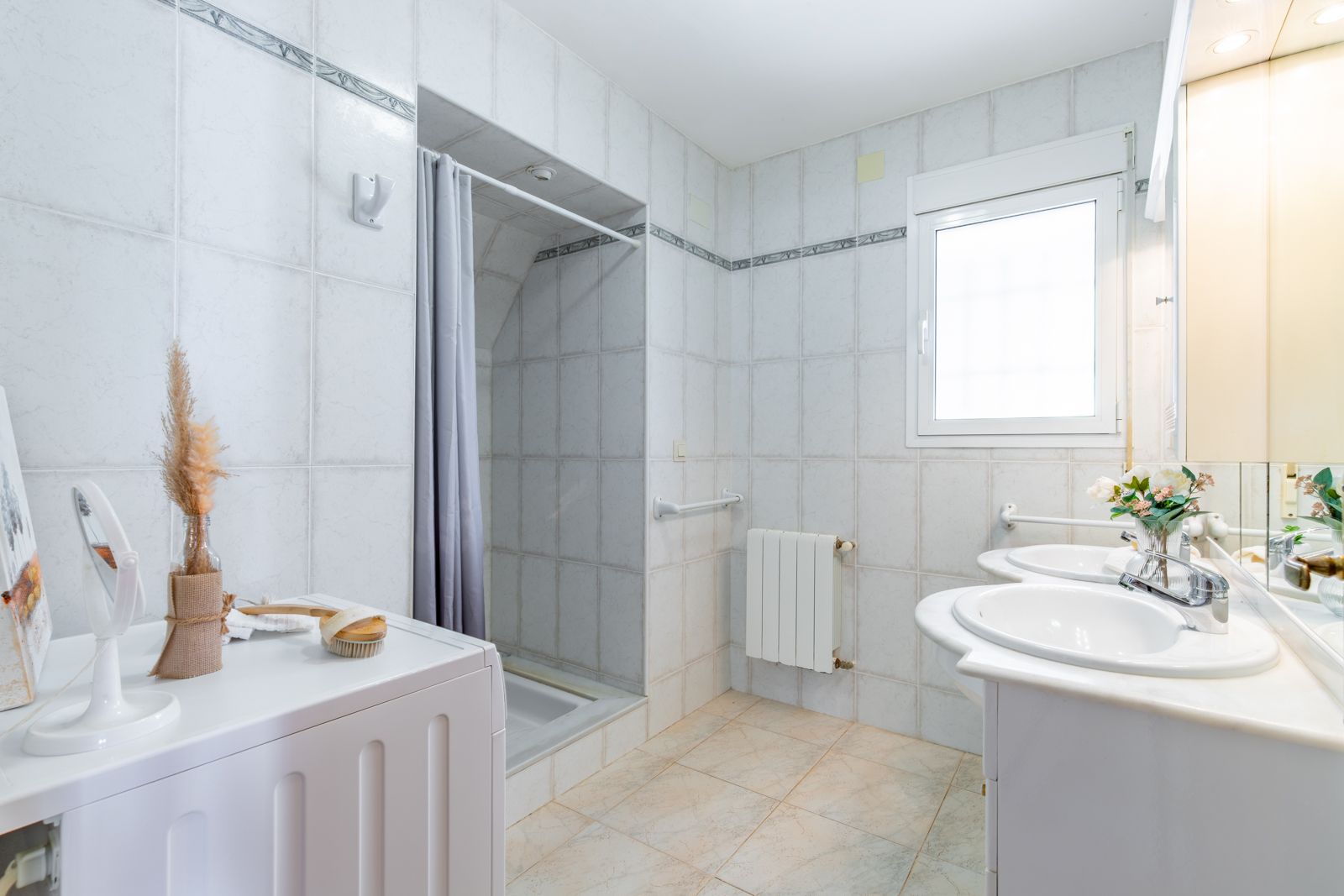 Funktionales Badezimmer mit Doppelwaschtisch aus Marmor, Waschmaschinenanschluss, begehbarer Dusche und Heizkörper unter dem Fenster.