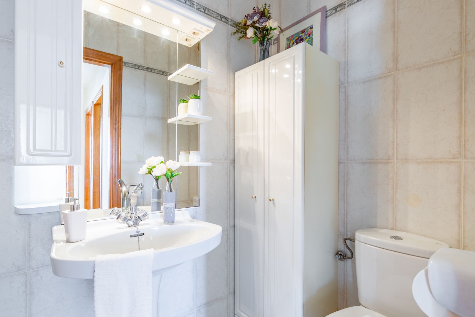 Funktionales Badezimmer mit Säulenwaschbecken, großem Spiegel mit integrierten Strahlern und Glasregalen sowie einem weißen Schrank vor neutralen Fliesen.