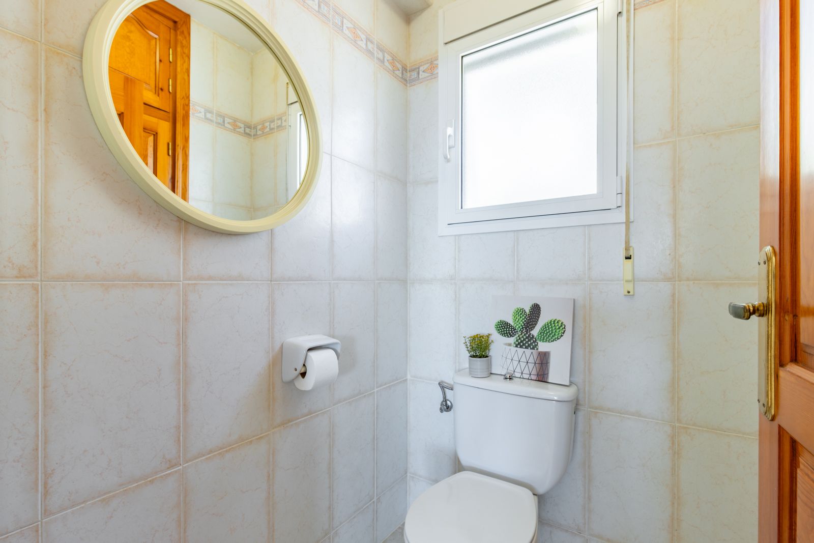 Funktionales Gäste-WC mit beigen Wandfliesen, einem runden Spiegel mit hellem Rahmen und einem Milchglasfenster für natürliches Tageslicht.