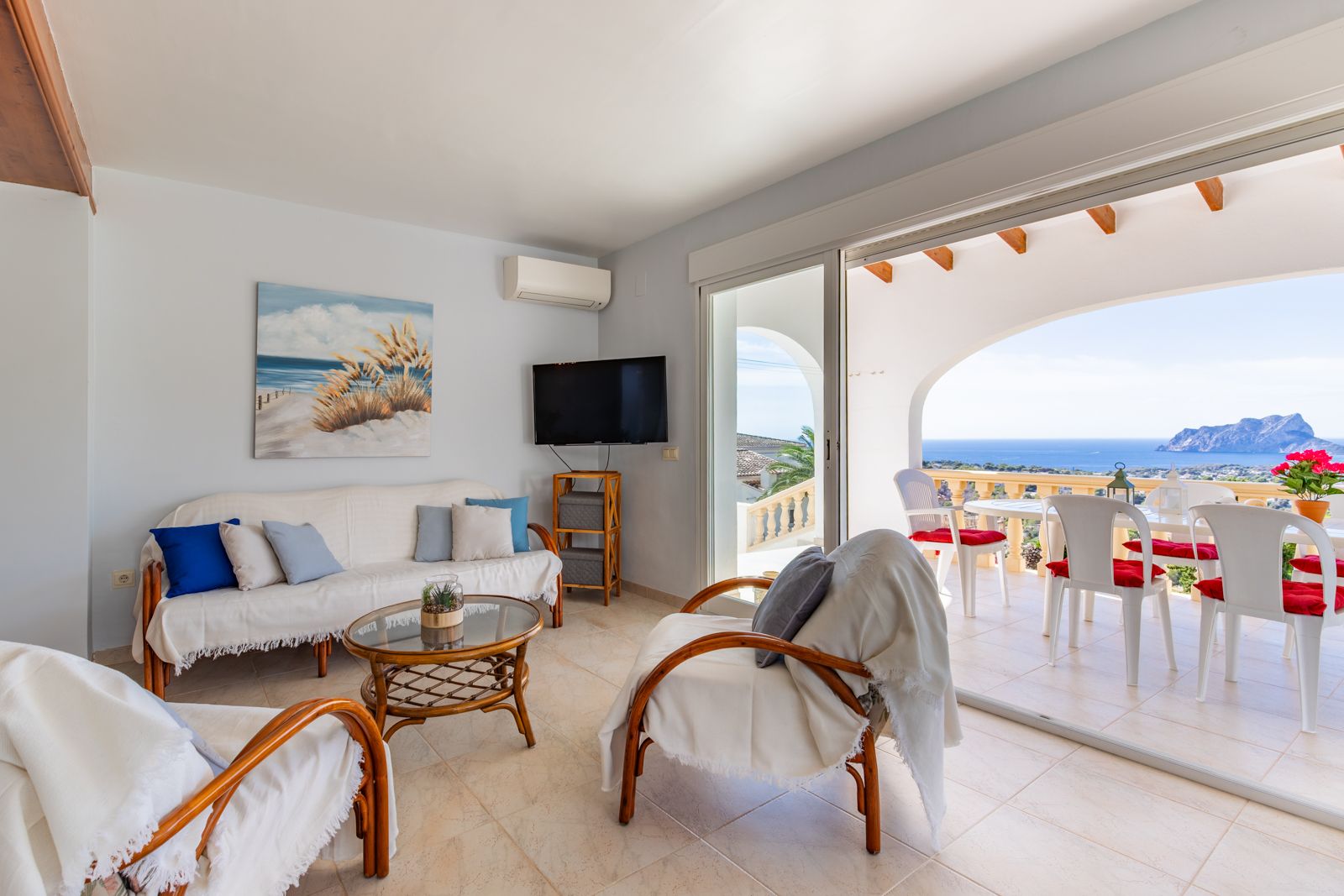 Heller Wohnbereich mit Rattanmöbeln und direktem Zugang zur überdachten Terrasse. Genießen Sie den Panoramablick auf das Mittelmeer und den Peñón de Ifach.