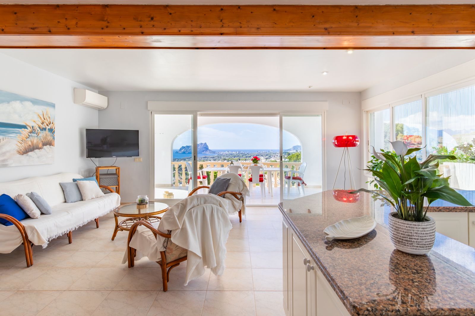 Heller Wohnbereich mit Sichtbalken aus Holz, Granit-Kücheninsel und Glasschiebetüren zur Terrasse mit Panoramablick auf das Mittelmeer und den Peñón de Ifach.