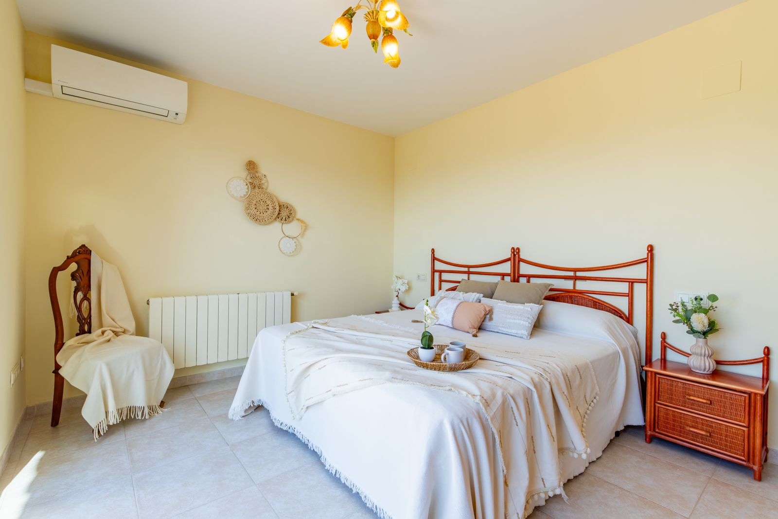 Helles Schlafzimmer mit Doppelbett, geschnitztem Holz-Kopfteil, passendem Nachttisch, Split-Klimaanlage, Heizkörper und neutralen Fliesenböden.