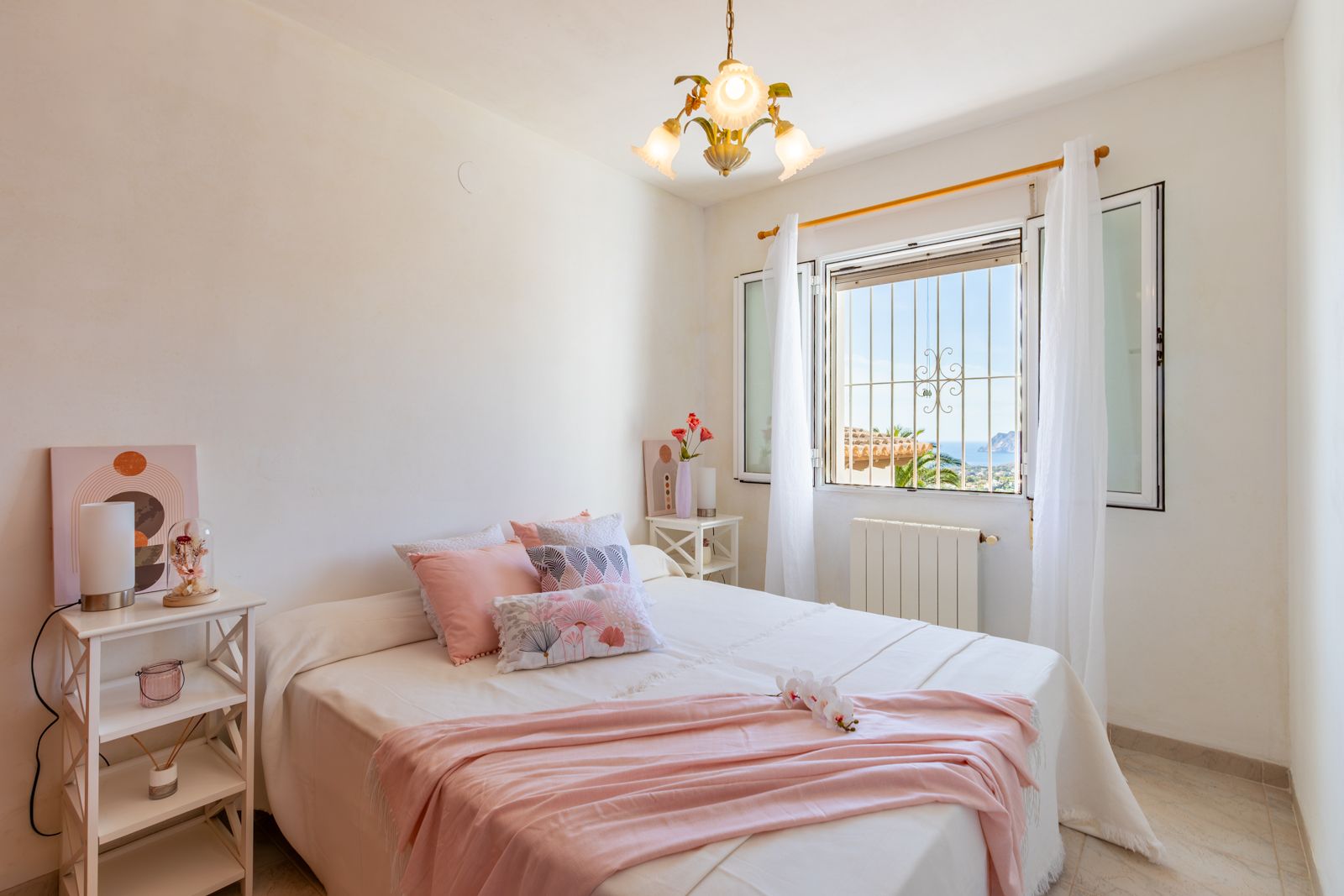 Helles Schlafzimmer mit Doppelbett, weißen Wänden und Fliesenboden. Das Fenster bietet einen weiten Blick auf das Mittelmeer und den markanten Peñón de Ifach.