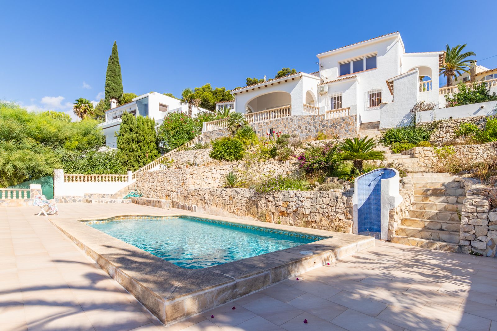 Privater Pool mit weitläufiger Sonnenterrasse, umgeben von terrassierten Gärten mit Natursteinmauern und einer Außendusche vor der weißen mediterranen Villa.