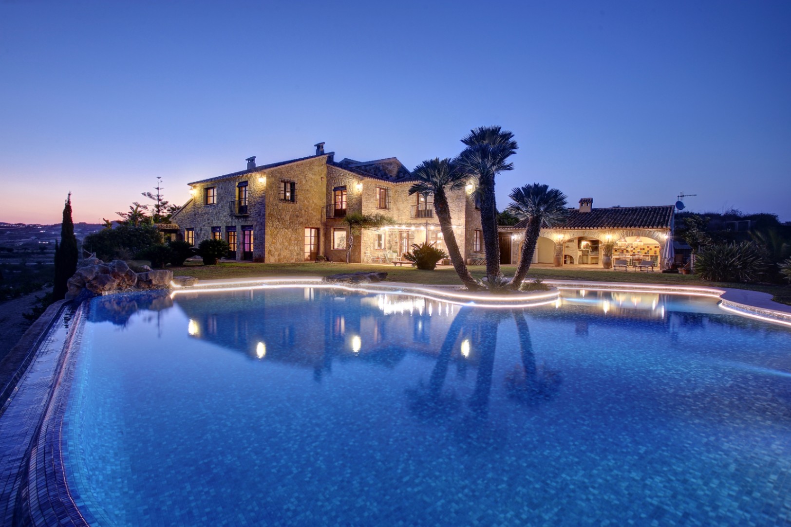 Abendliche Außenansicht der Natursteinfassade und des organisch geformten Pools mit LED-Beleuchtung sowie der angrenzenden Außenküche mit Grillbereich.