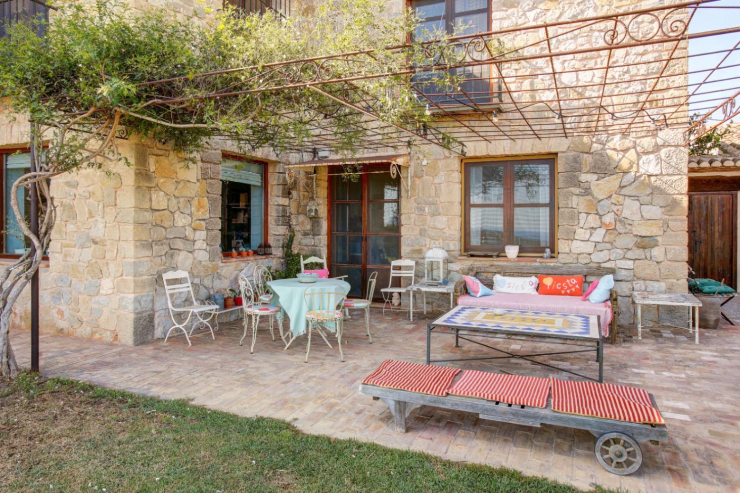 Außenterrasse mit Natursteinfassade, Terrakottaboden und einer schmiedeeisernen Pergola. Der Bereich verfügt über rustikale Sitzgelegenheiten und einen Couchtisch mit Mosaikplatte.