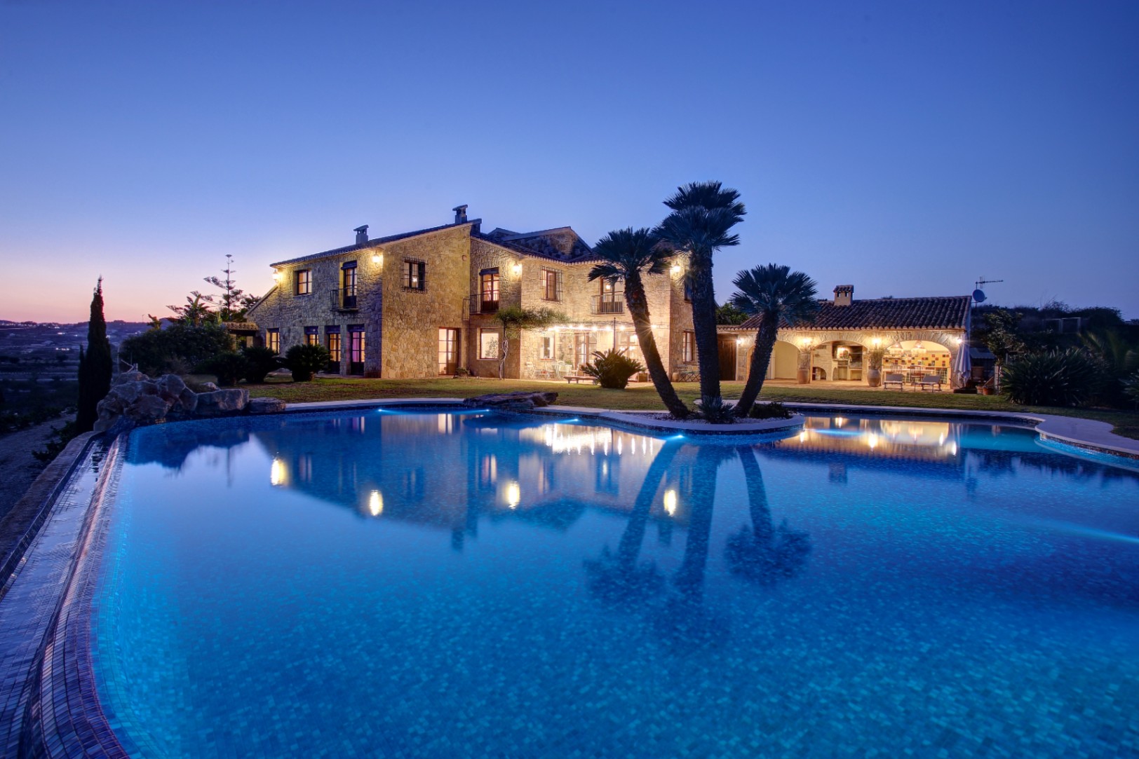 Dämmerungsansicht der Steinfassade und des beleuchteten Pools mit Außenküche und in die mediterrane Gartenanlage integrierten Palmen.