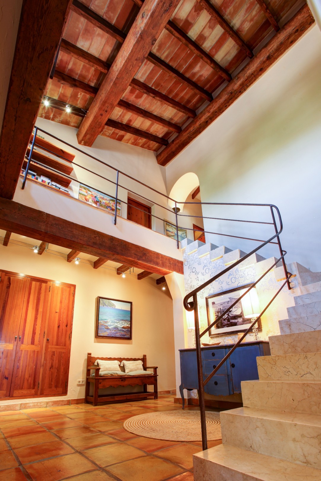 Eingangshalle mit doppelter Deckenhöhe, sichtbaren Holzbalken und Terrakotta-Elementen. Eine massive Marmortreppe mit Schmiedeeisengeländer führt zur oberen Galerie.