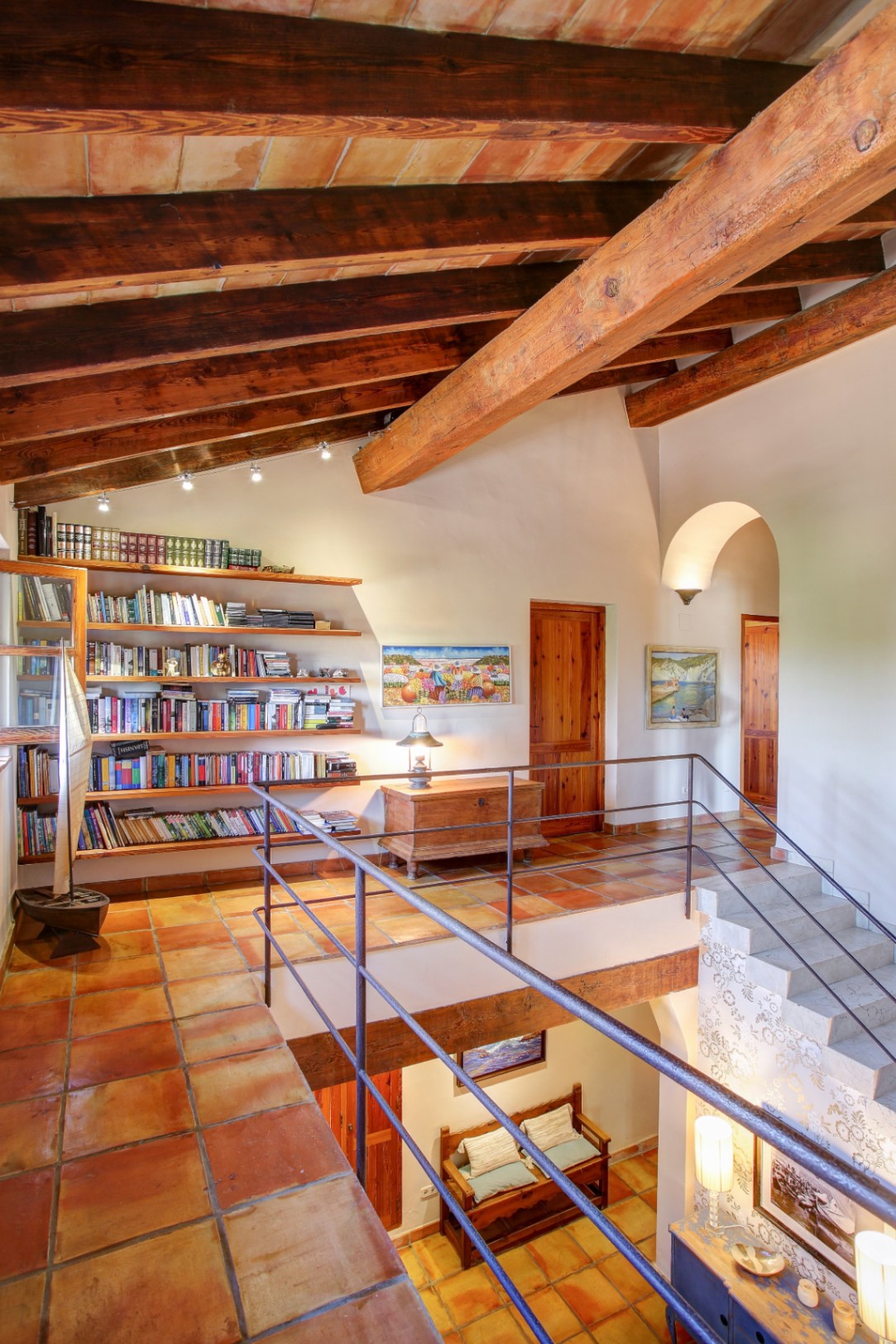 Flur im Obergeschoss mit sichtbaren Holzbalken, Terrakottaboden, integriertem Bücherregal und Blick auf den darunter liegenden Wohnbereich durch die Galerie.