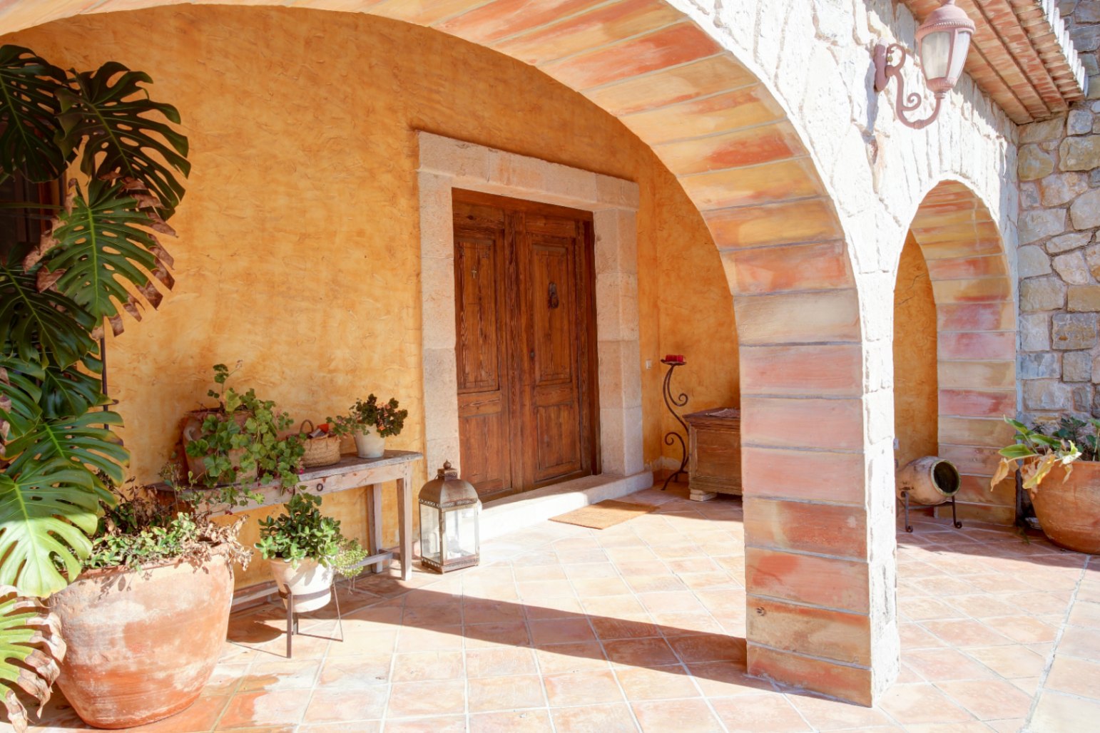 Haupteingang unter einem rustikalen Steinbogen-Porch mit Terrakottaboden, einer massiven Holztür und üppiger mediterraner Topfbepflanzung.