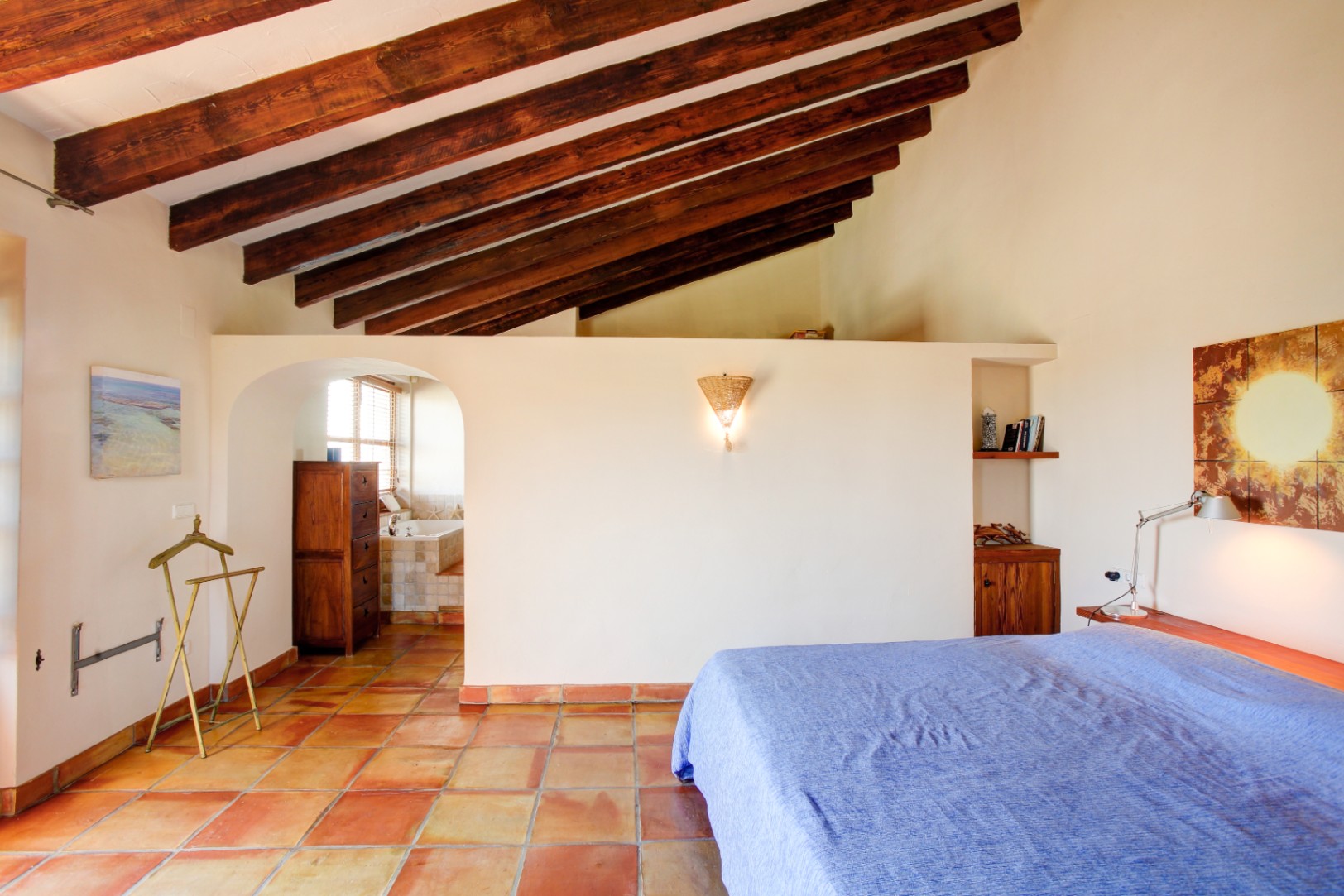 Hauptschlafzimmer mit hohen Decken und sichtbaren Holzbalken, traditionellen Terrakotta-Fliesen und einem Rundbogen-Durchgang zum integrierten Badezimmer.