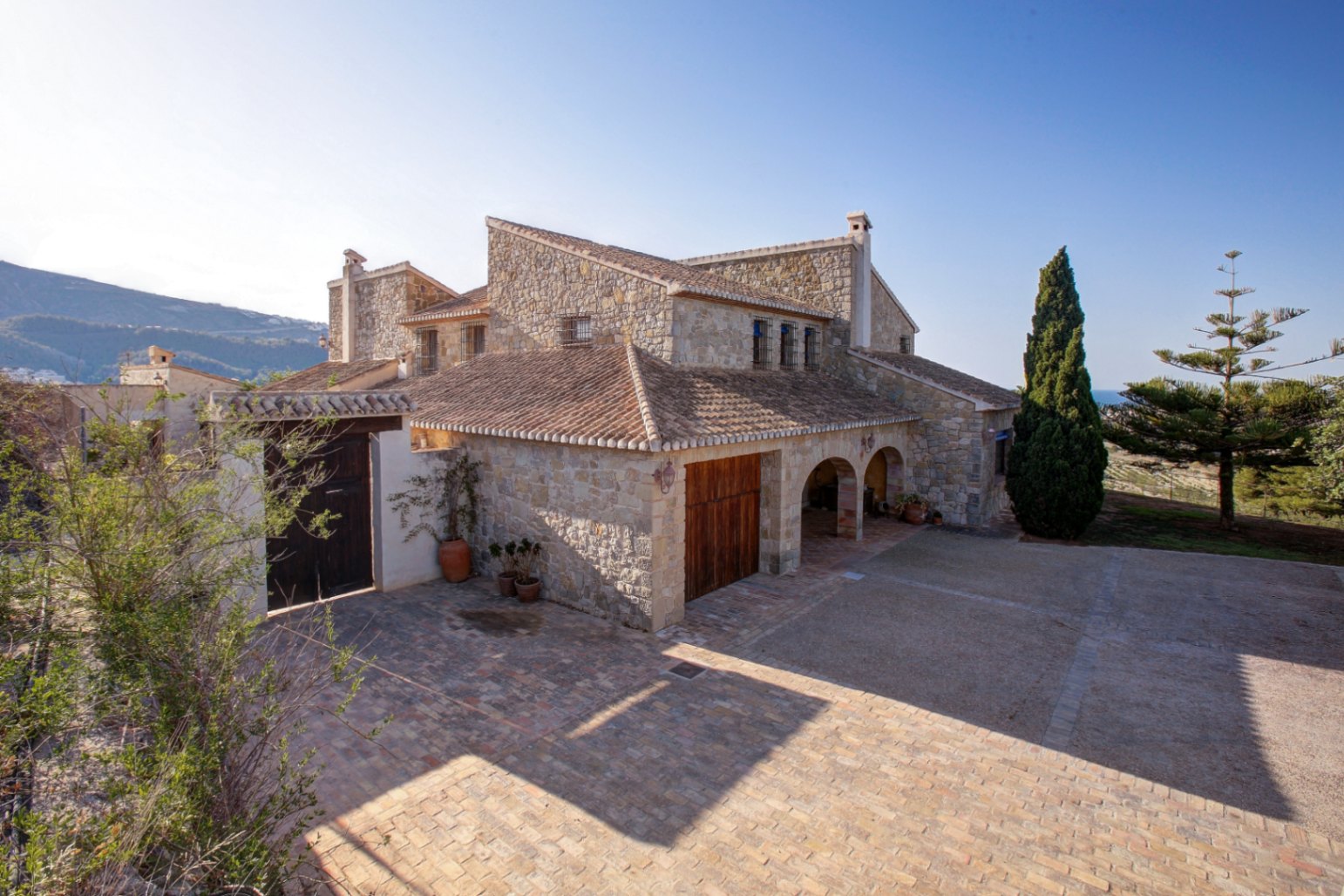 Natursteinfassade mit traditionellen Rundbögen, Tonziegeldach und weitläufigem, mit Terrakotta gepflastertem Innenhof unter mediterraner Sonne.