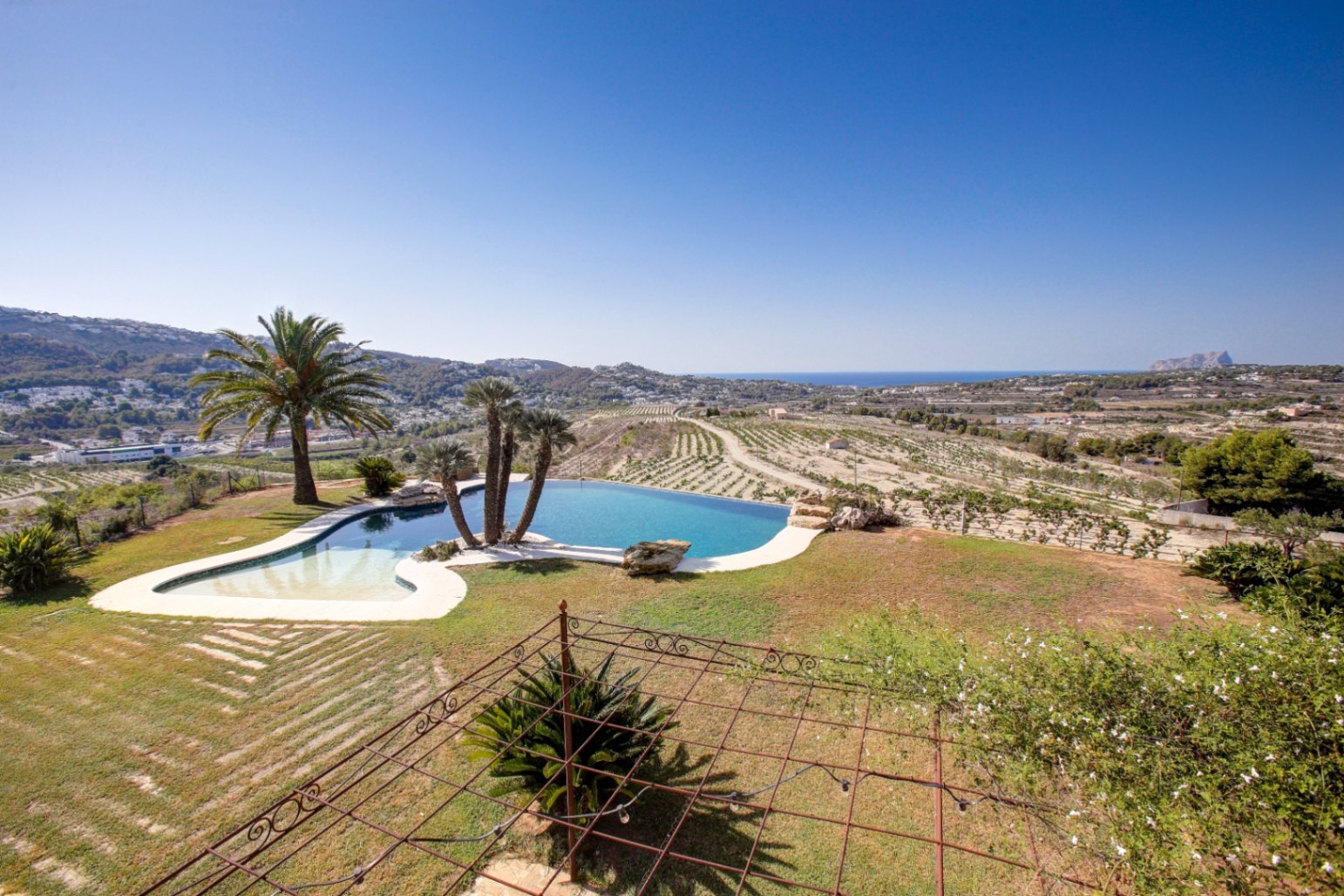 Organisch geformter Infinity-Pool mit weitem Blick auf das Mittelmeer und den Peñón de Ifach, umgeben von weitläufigen Gartenanlagen und Weinreben.