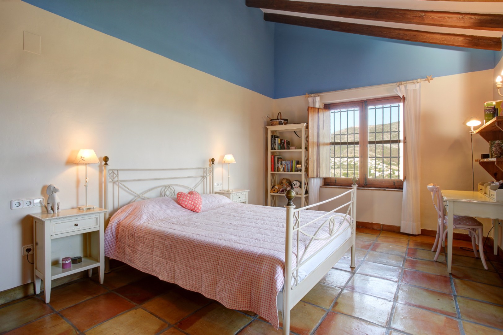 Rustikales Schlafzimmer mit Sichtbalkendecke, Terrakottaboden und weißem Eisenbett. Es bietet einen Arbeitsbereich und weiten Blick auf die umliegende Hügellandschaft.
