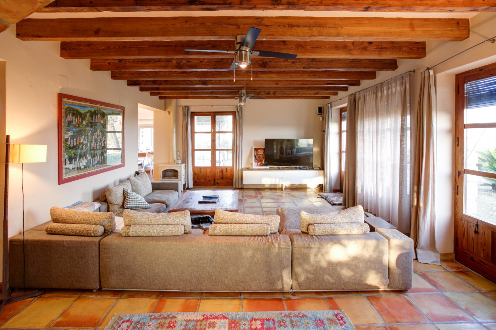 Rustikales Wohnzimmer mit sichtbaren Holzbalken, Terrakottaboden, einem weißen Holzofen und großen Fenstertüren aus Holz mit direktem Zugang zum Garten.