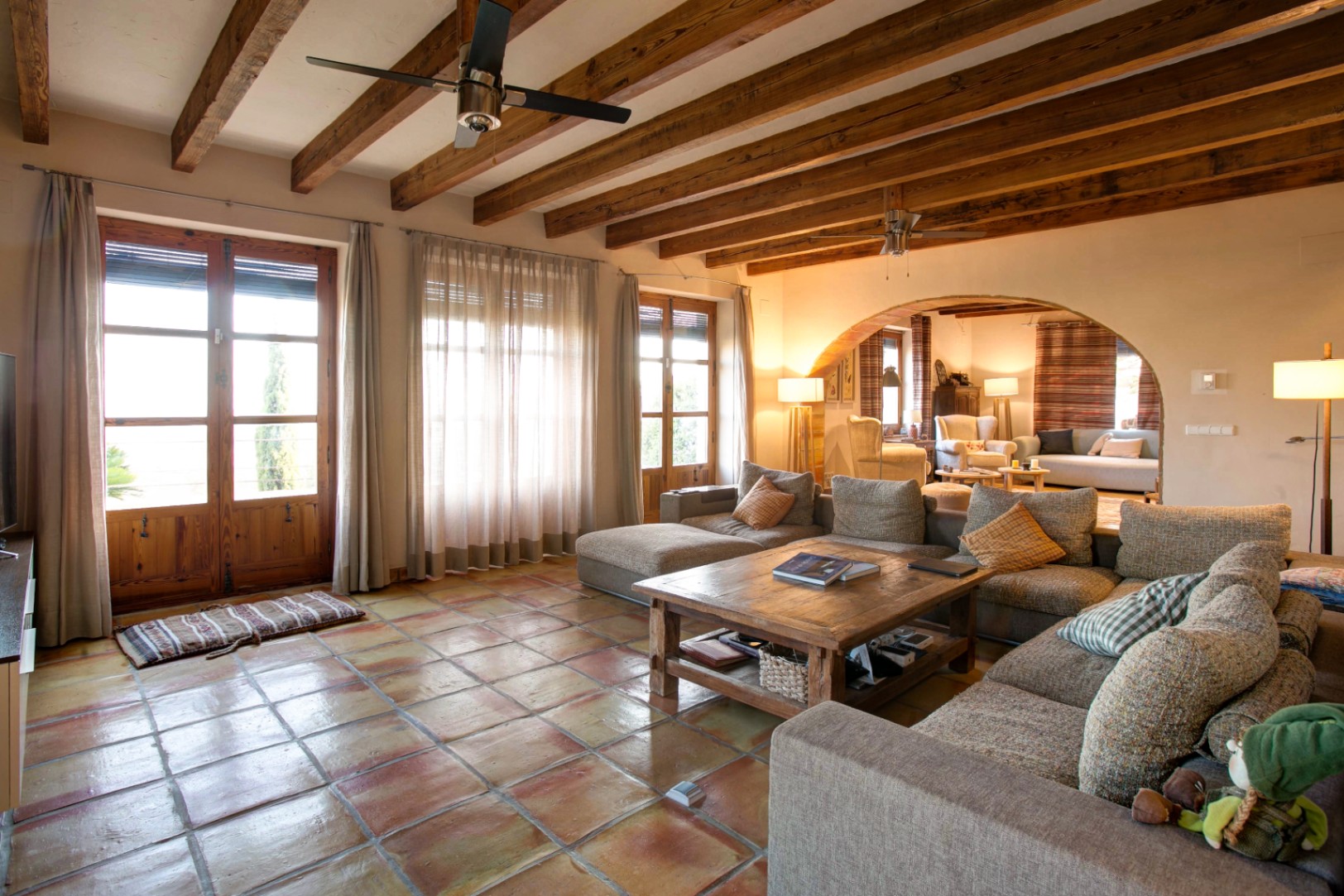 Rustikales Wohnzimmer mit Terrakottaboden, sichtbaren Holzbalkendecken und einem Rundbogen. Große Fenstertüren aus Holz sorgen für viel natürliches Licht im Raum.